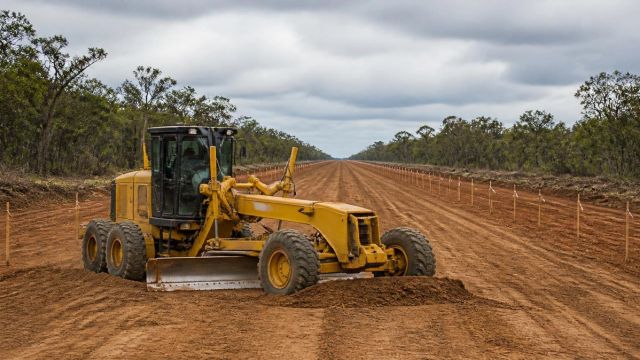 Cleared, graded highway corridor with a motor grader and rows of survey stakes receding into the distance.