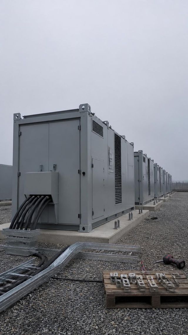 Rows of grey battery storage container units on a gravel industrial yard, nearest one unfinished with exposed cabling.