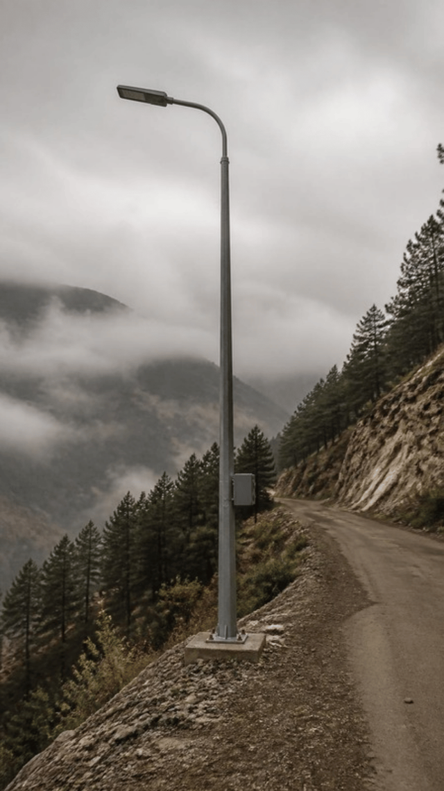 A galvanised steel highway lighting mast standing unlit on a concrete base at the edge of a hill road, with mist in the valley below.