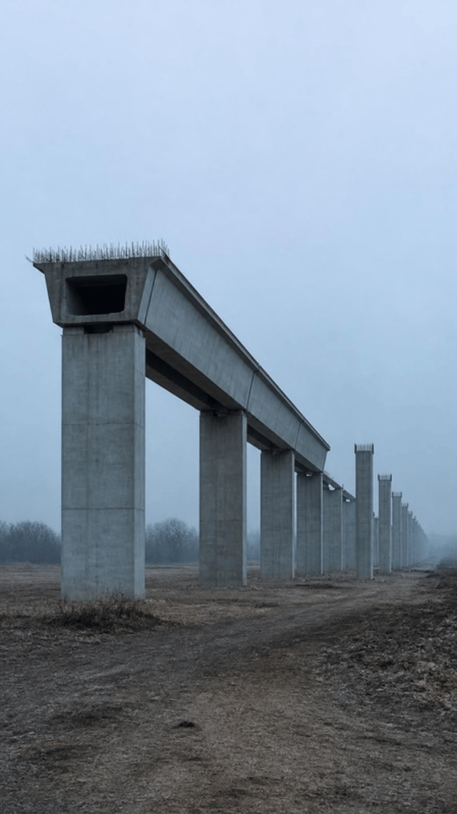 An unfinished elevated metro viaduct, with precast box girder segments joined into a deck that stops in mid air above two bare concrete piers.
