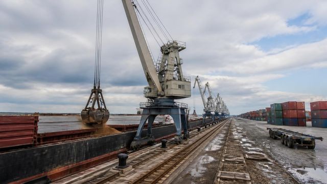 A harbour crane with its grab over a ship hold on a working quay, with rails and cargo stacks receding into the distance.