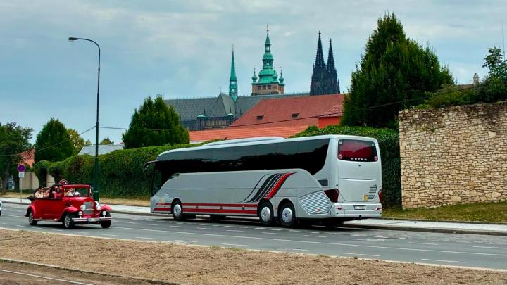 A silver and red coach bus parked on asphalt, with a church and trees in the background under a cloudy sky.