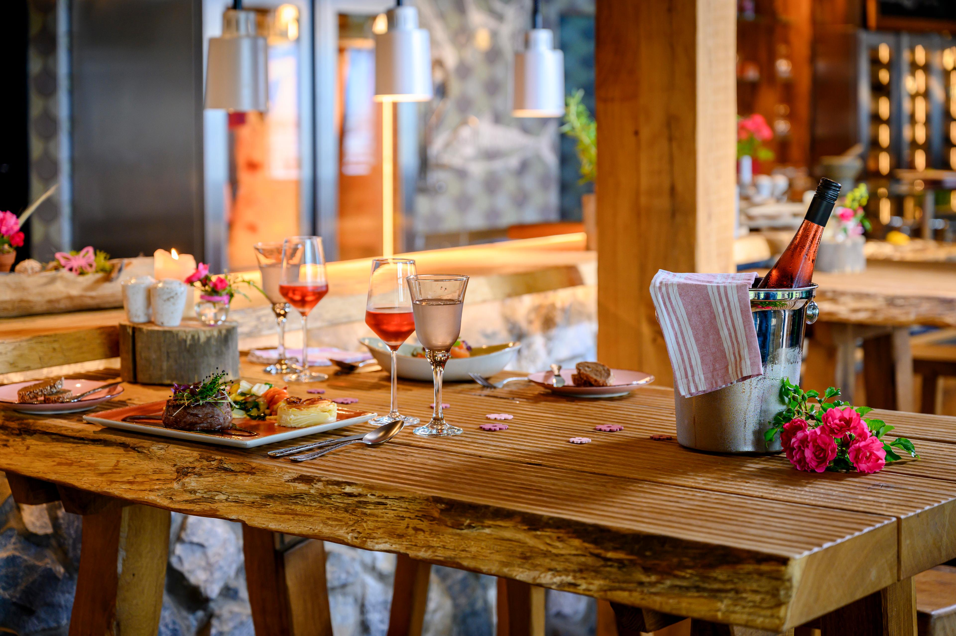 Ein rustikaler Holztisch in einem Restaurant, gedeckt mit Weingläsern, Tellern mit Gourmetgerichten und einem Weinkühler mit Roséwein.