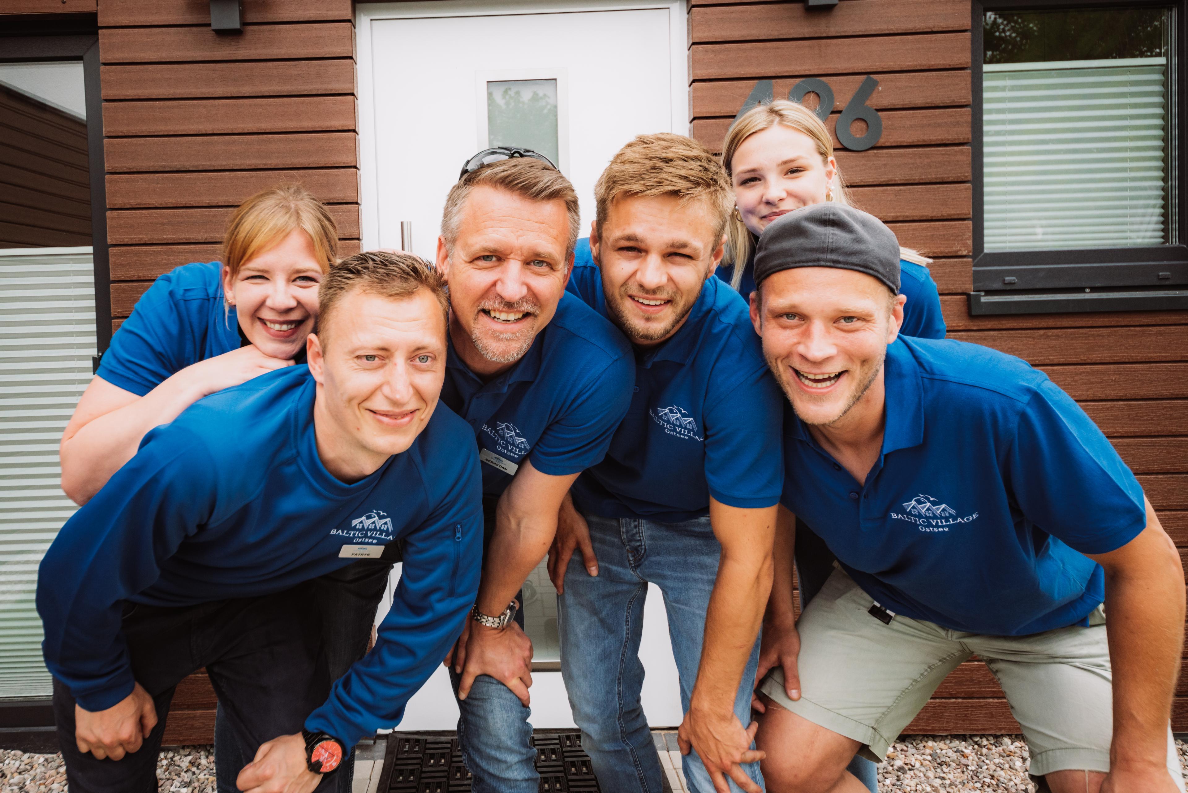 Eine Gruppe von sechs Personen in blauen Hemden posiert lächelnd vor einem Holzhaus