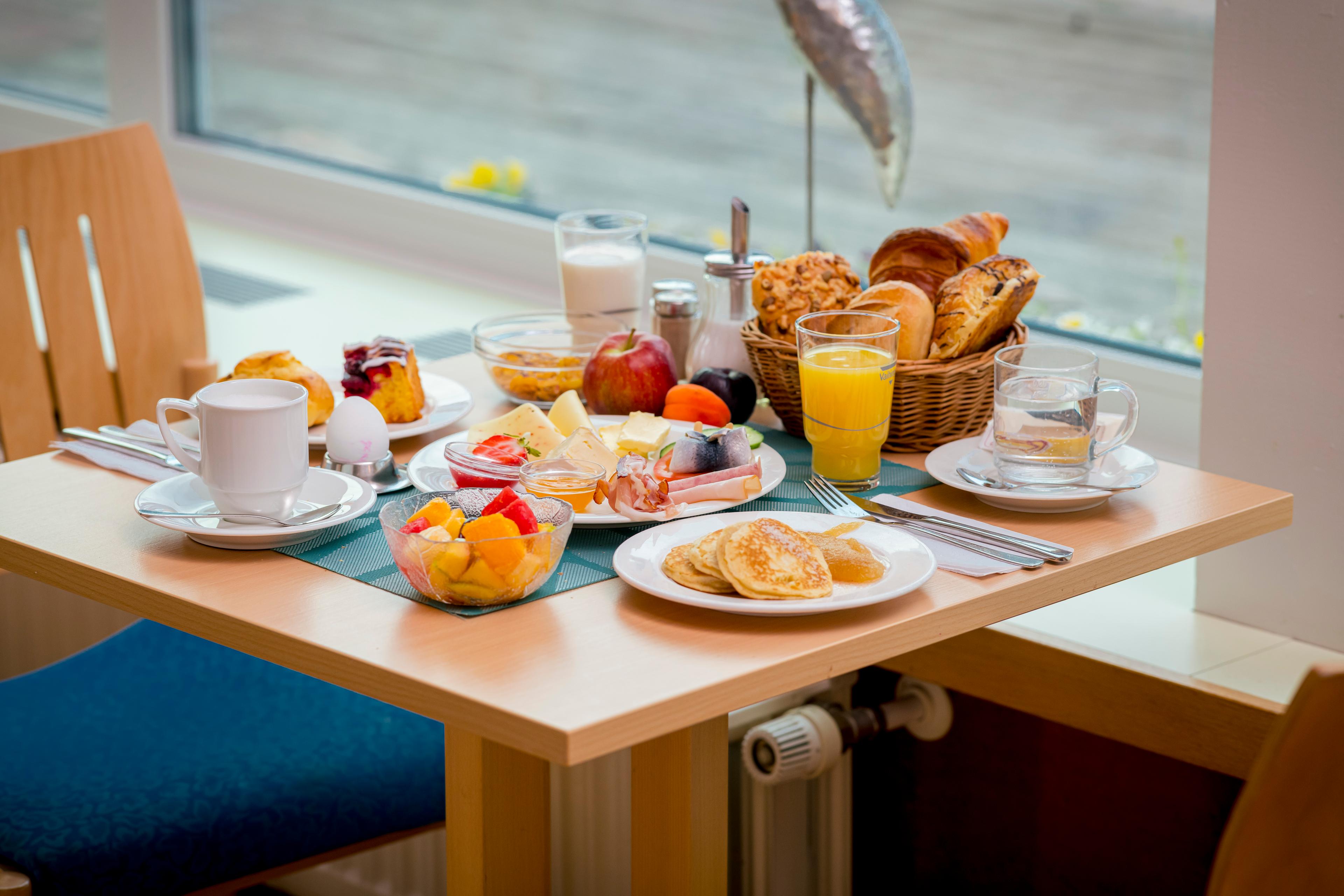 Ein reichhaltiges Frühstücksbuffet mit Obst, Gebäck, Käse, Aufschnitt und Getränken auf einem Tisch am Fenster