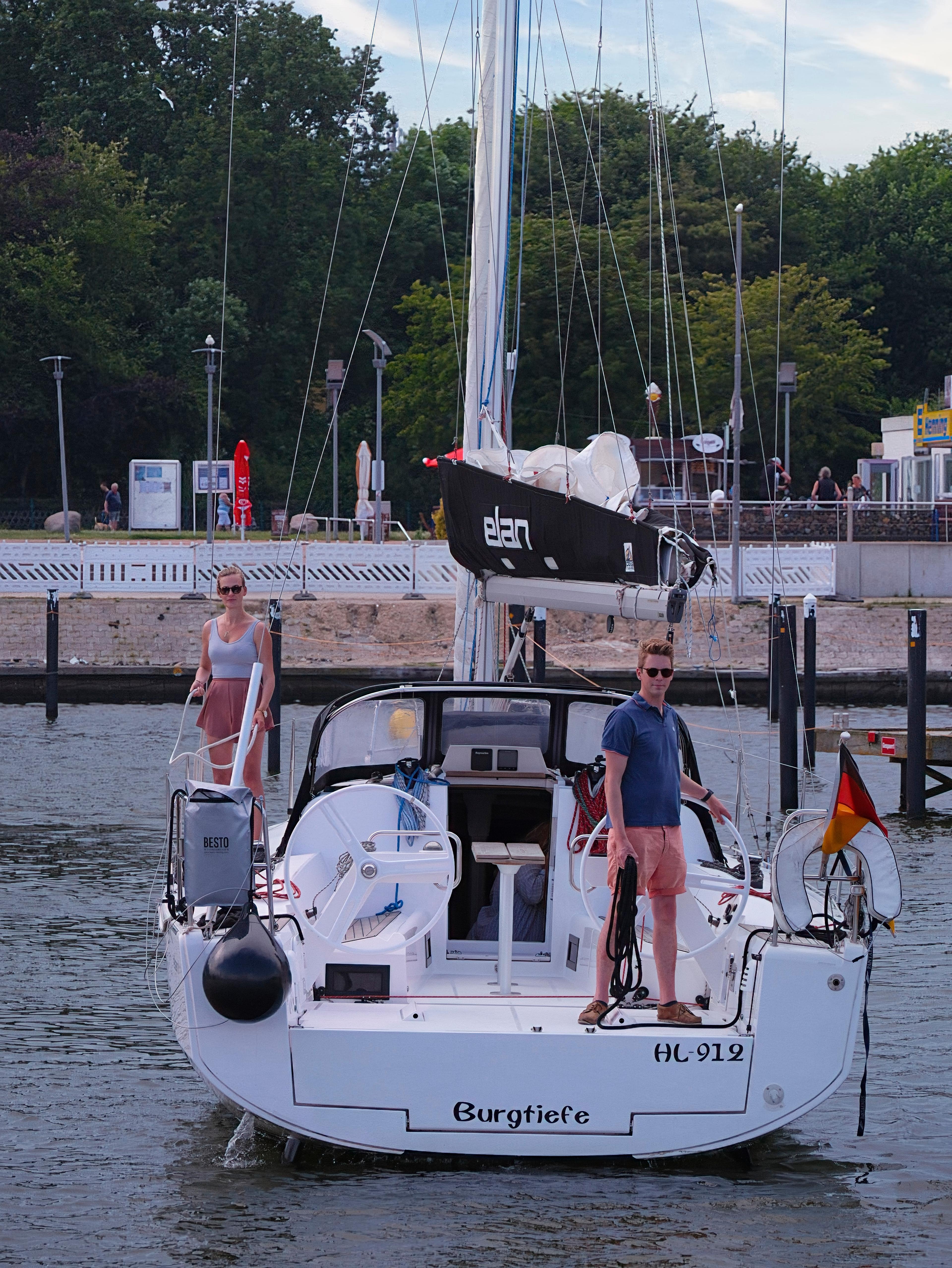 Ein Segelboot mit zwei Personen im Hafen, umgeben von Bäumen und Gebäuden