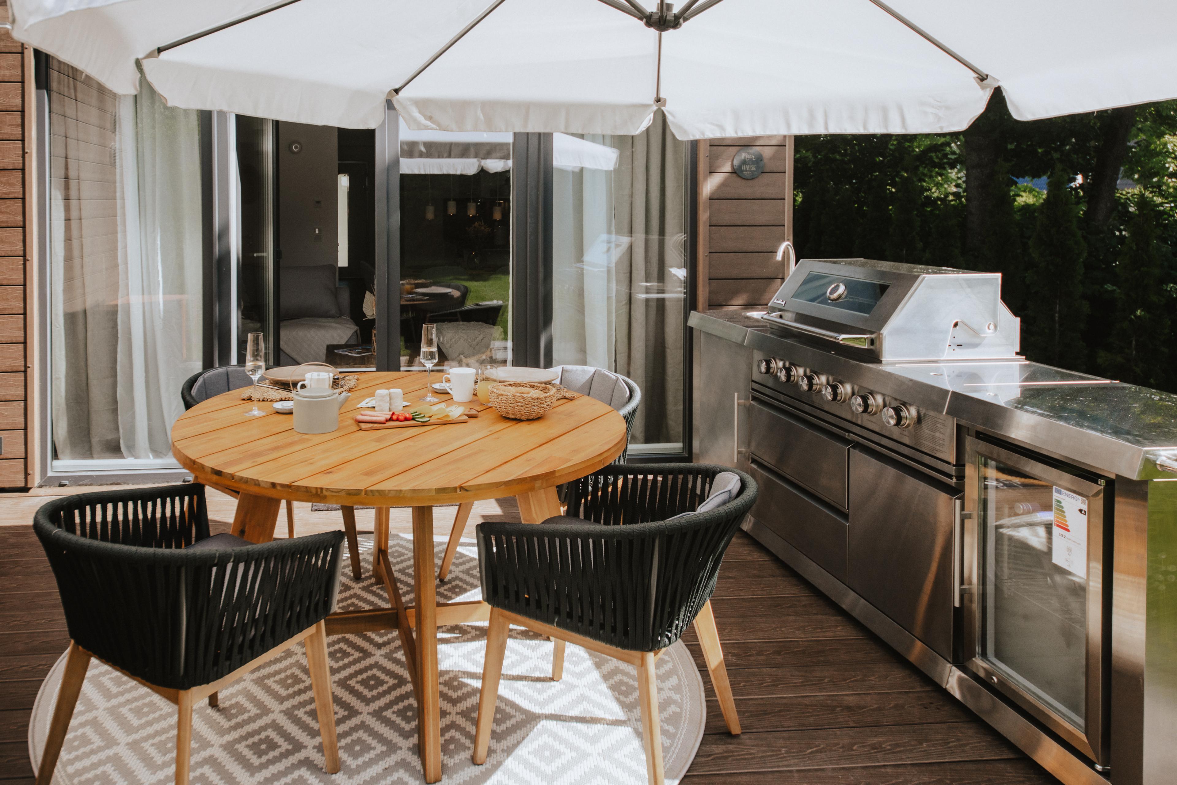 Eine moderne Terrasse mit einem runden Holztisch, vier Stühlen und einem großen Grill unter einem Sonnenschirm