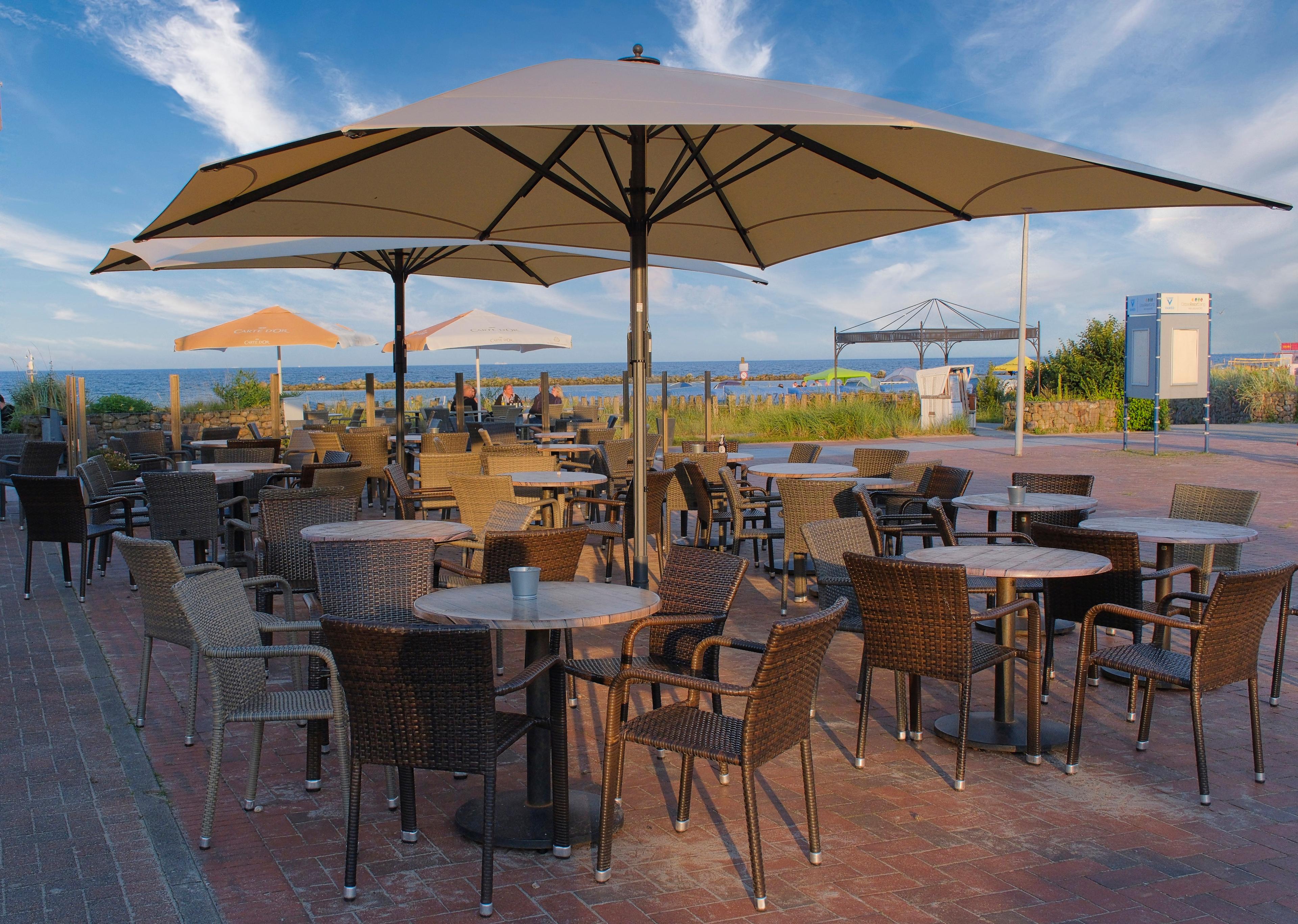 Ein Strandcafé mit leeren Tischen und Stühlen unter großen Sonnenschirmen, mit Blick auf das Meer im Hintergrund