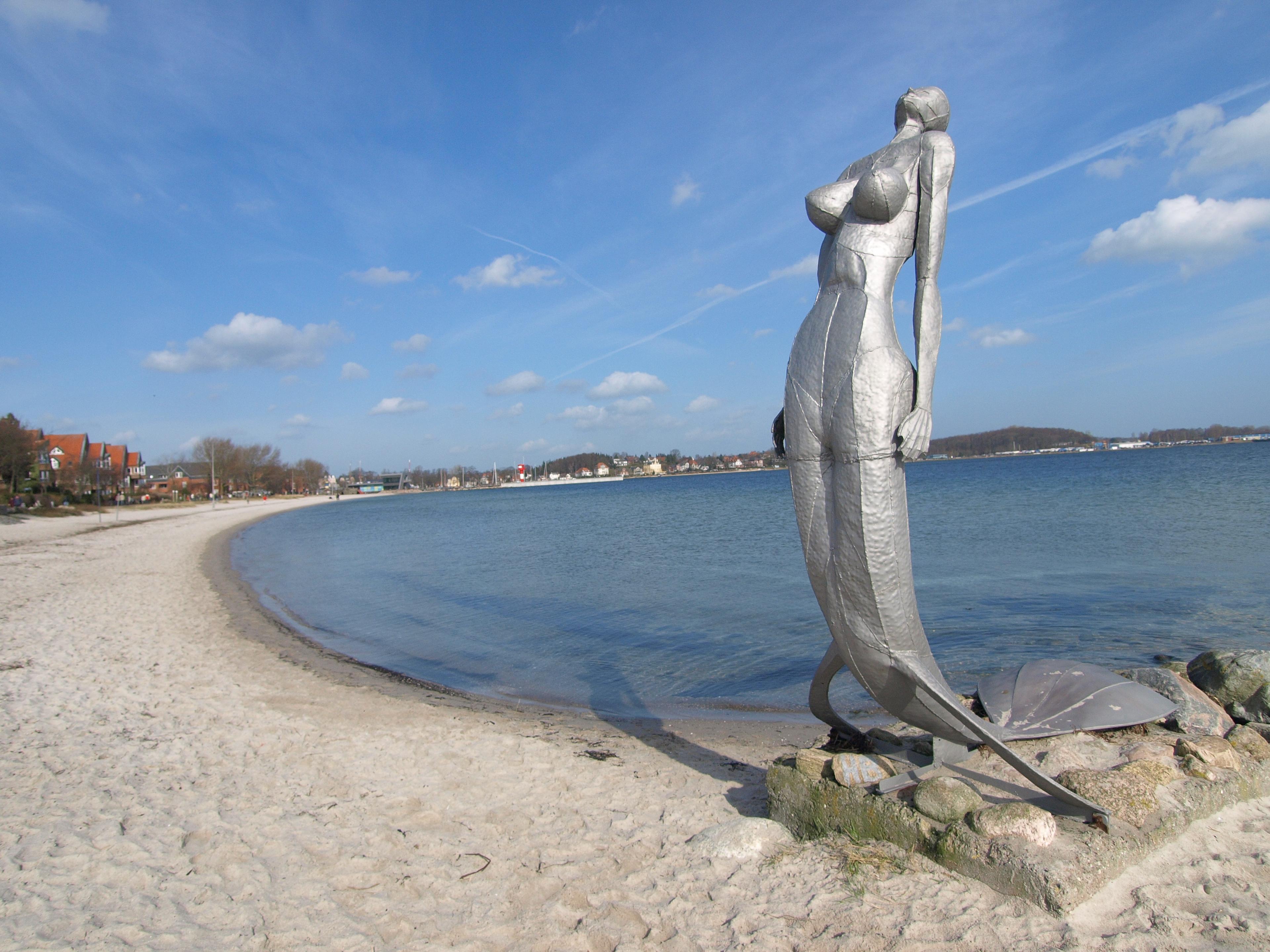 Eine große Metallstatue am Strand mit Blick auf das Meer und einen klaren blauen Himmel