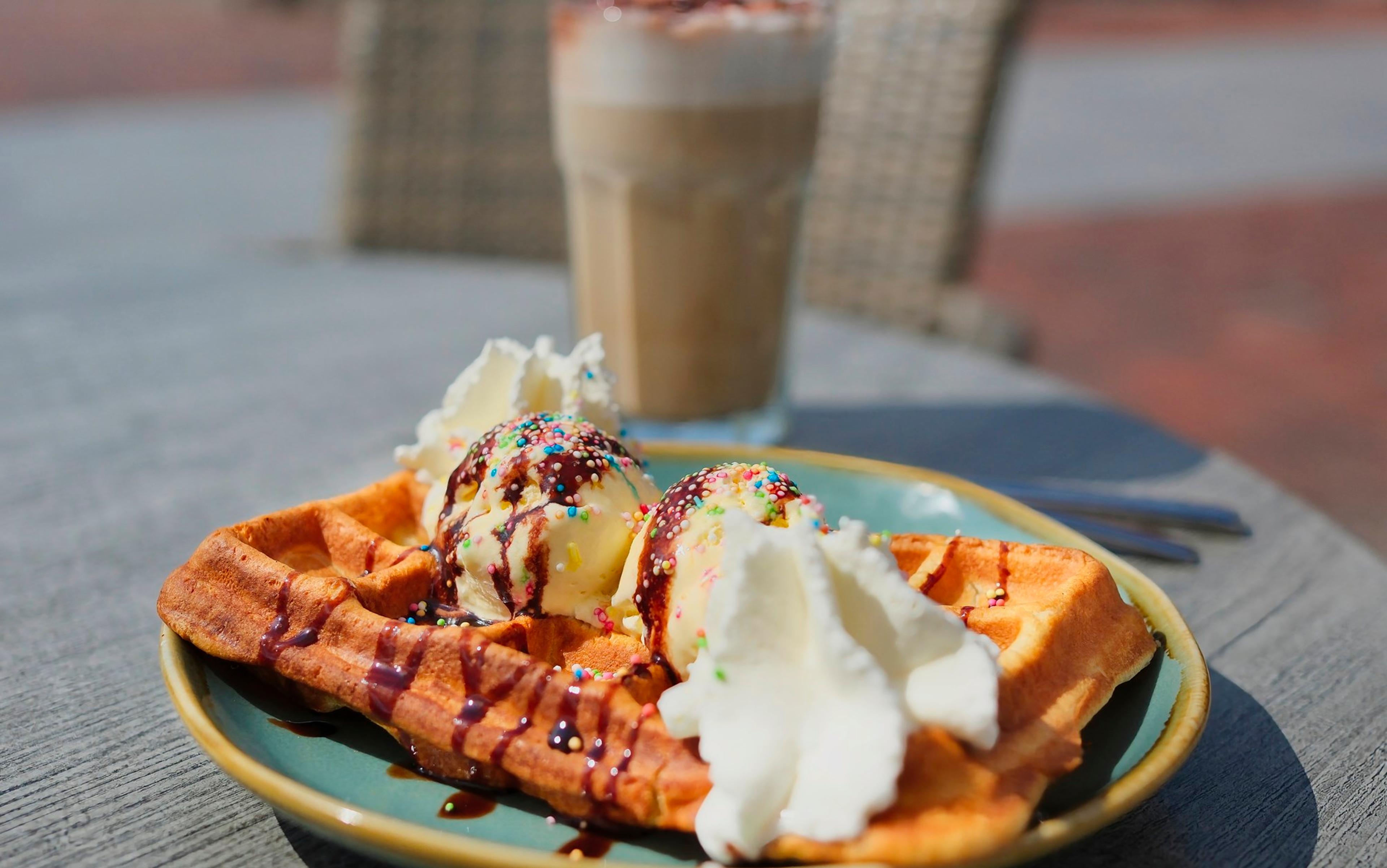 Belgische Waffeln mit zwei Kugeln Vanilleeis, Schokoladensauce und Sahne, serviert auf einem Teller mit einem Glas Eiskaffee im Hintergrund