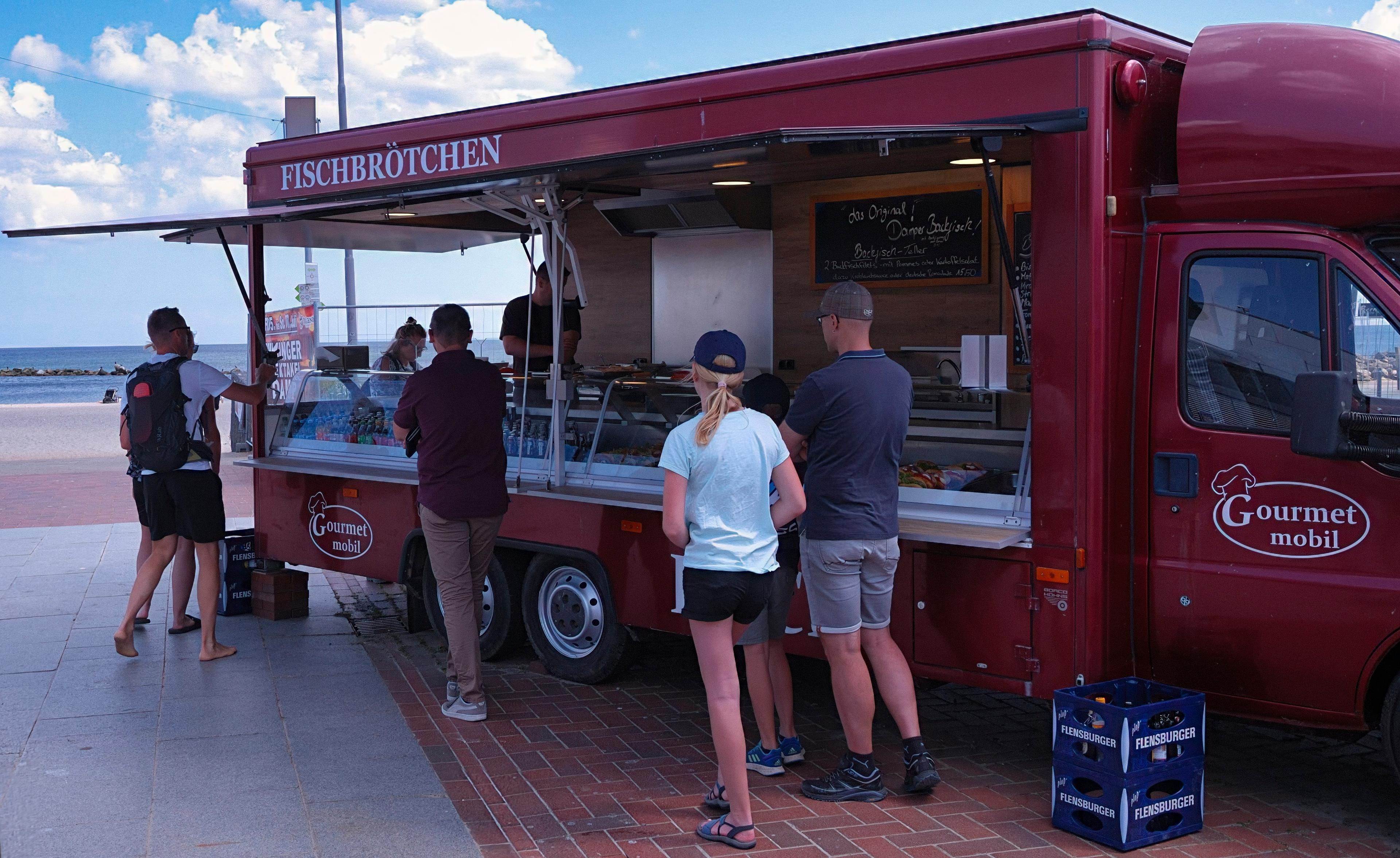 Ein roter Food Truck mit der Aufschrift 'Fischbrötchen' steht am Strand, umgeben von Kunden.