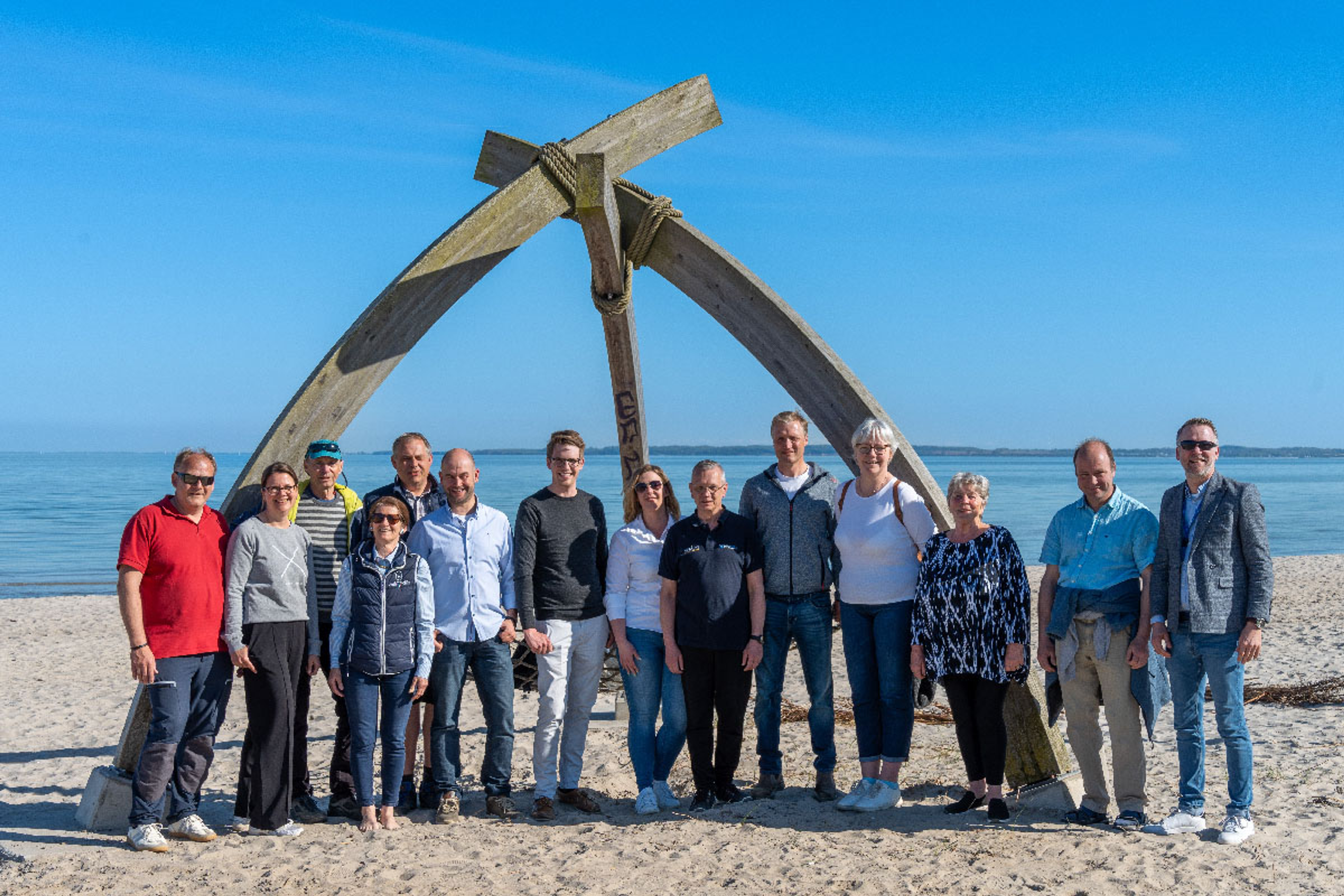 Eine Gruppe von Menschen steht vor einer großen Holzskulptur am Strand mit Meer im Hintergrund