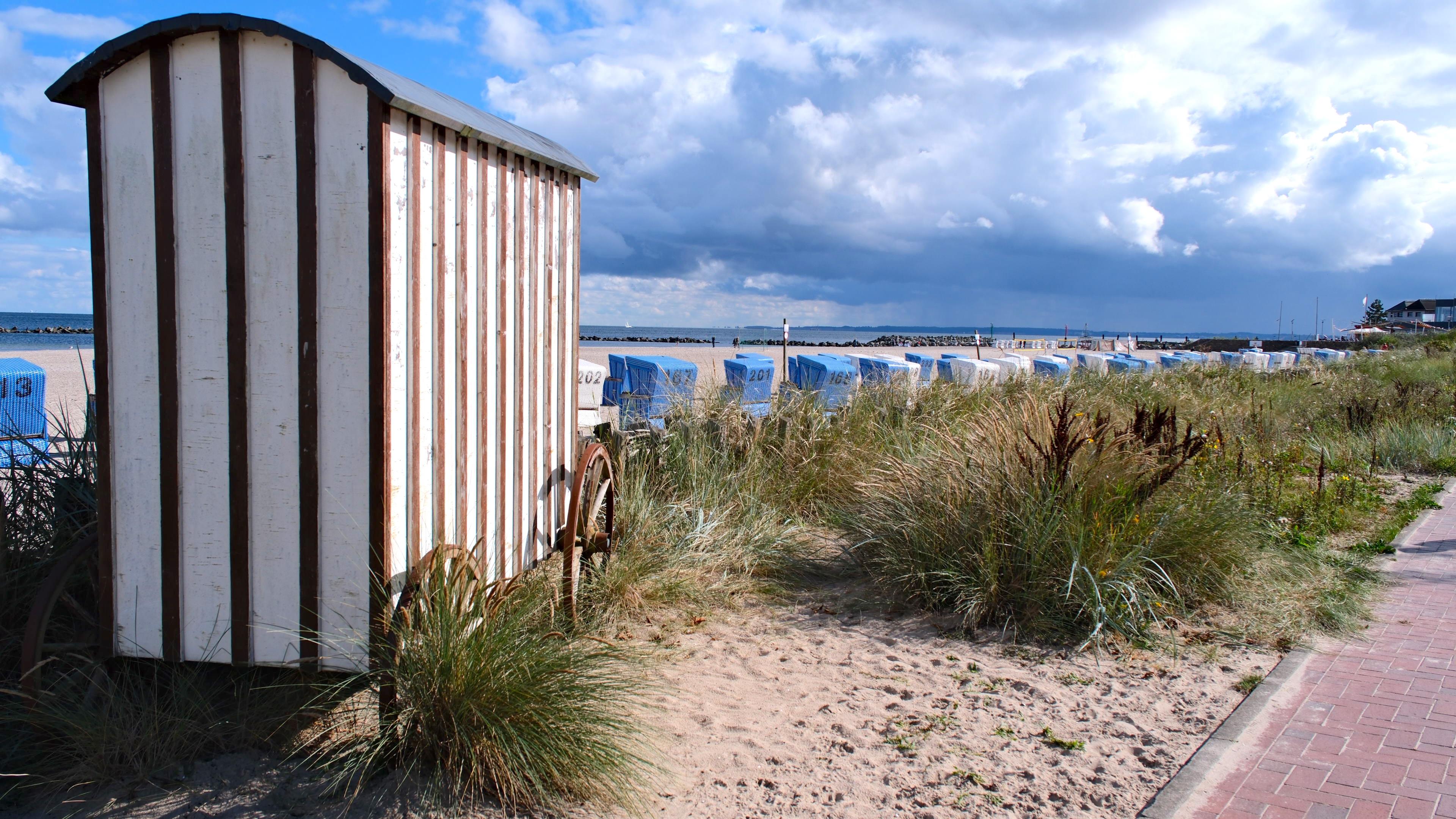 Eine historische Strandkabine neben einer Reihe von Strandkörben an einem sandigen Küstenabschnitt mit Gras und Wolken am Himmel