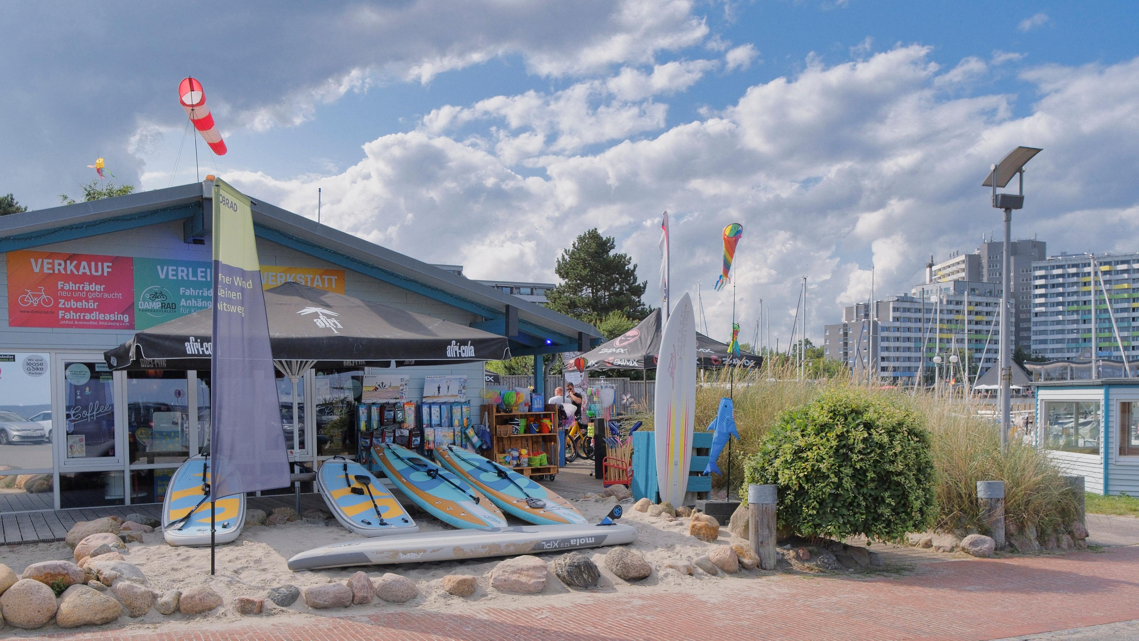 Ein Strandshop mit Surfboards, Fahrradverleih und bunten Fahnen vor einem bewölkten Himmel