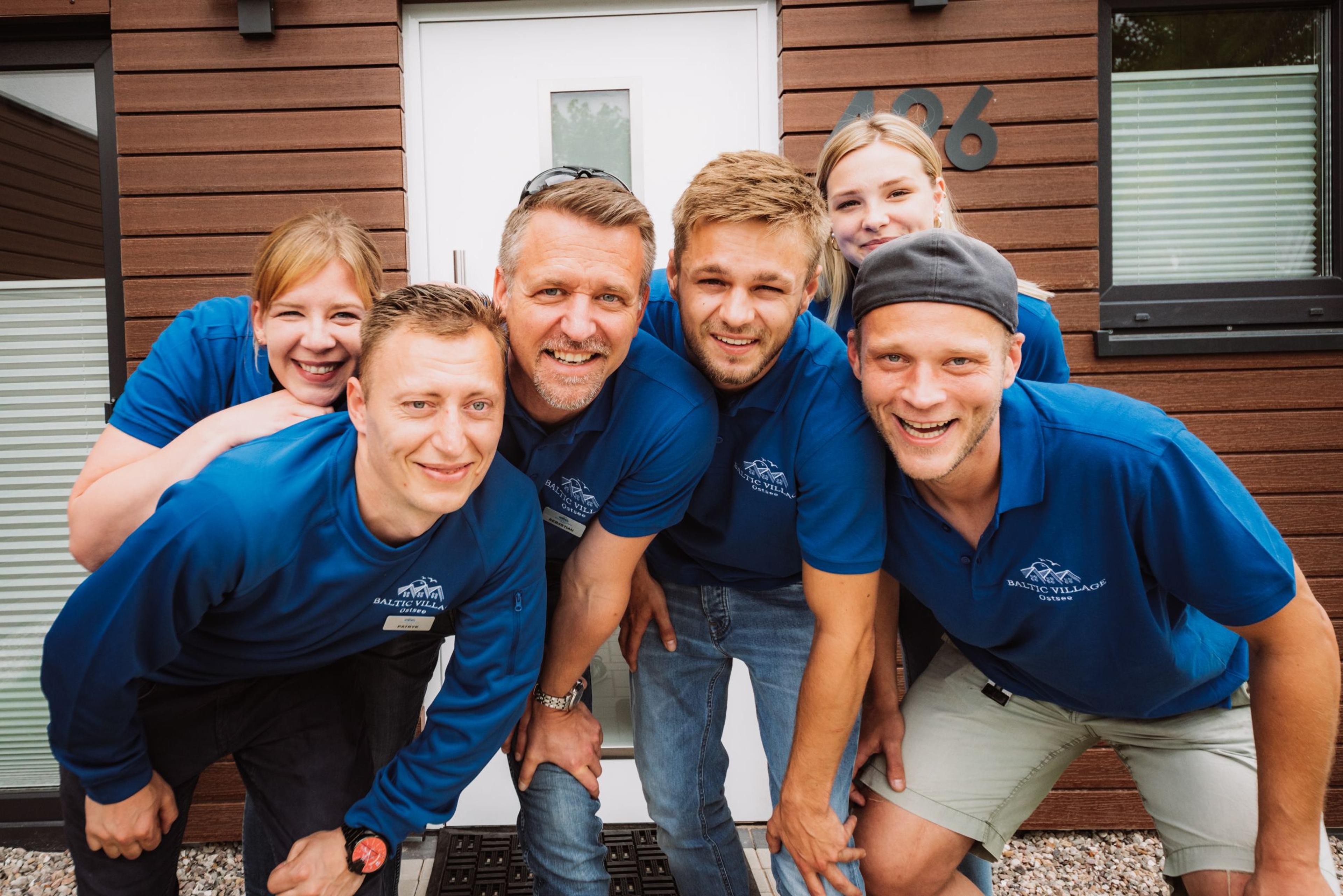 Eine Gruppe von sechs Personen in blauen Hemden posiert lächelnd vor einem modernen Holzhaus.