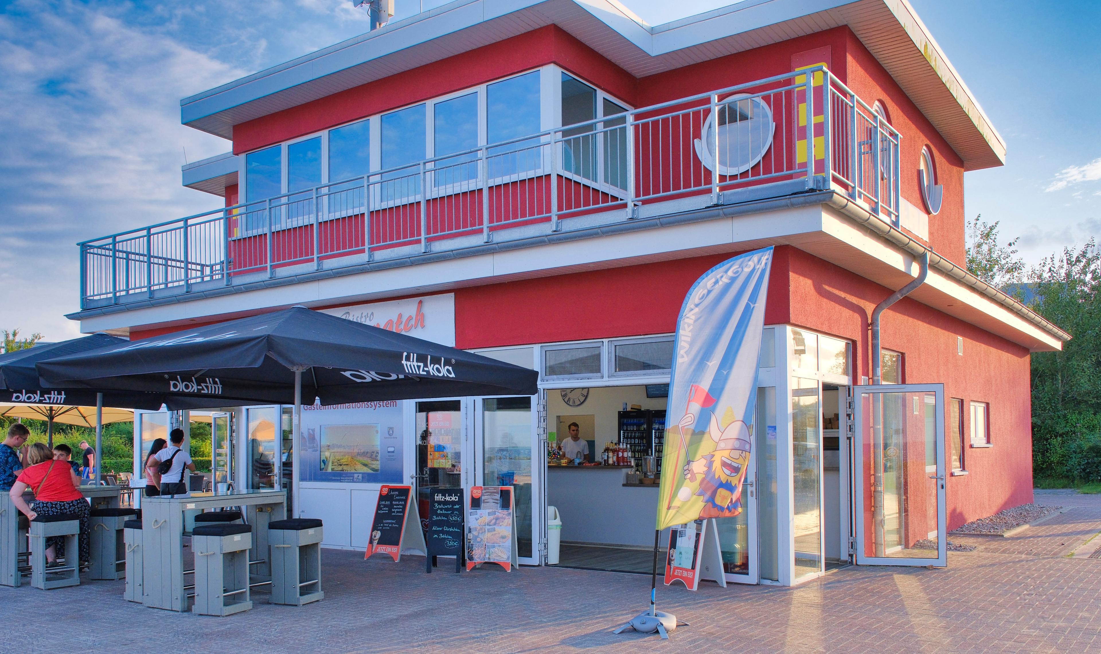 Ein modernes Café mit roter Fassade, großen Fenstern und einem Außenbereich mit Sonnenschirmen und Sitzgelegenheiten.