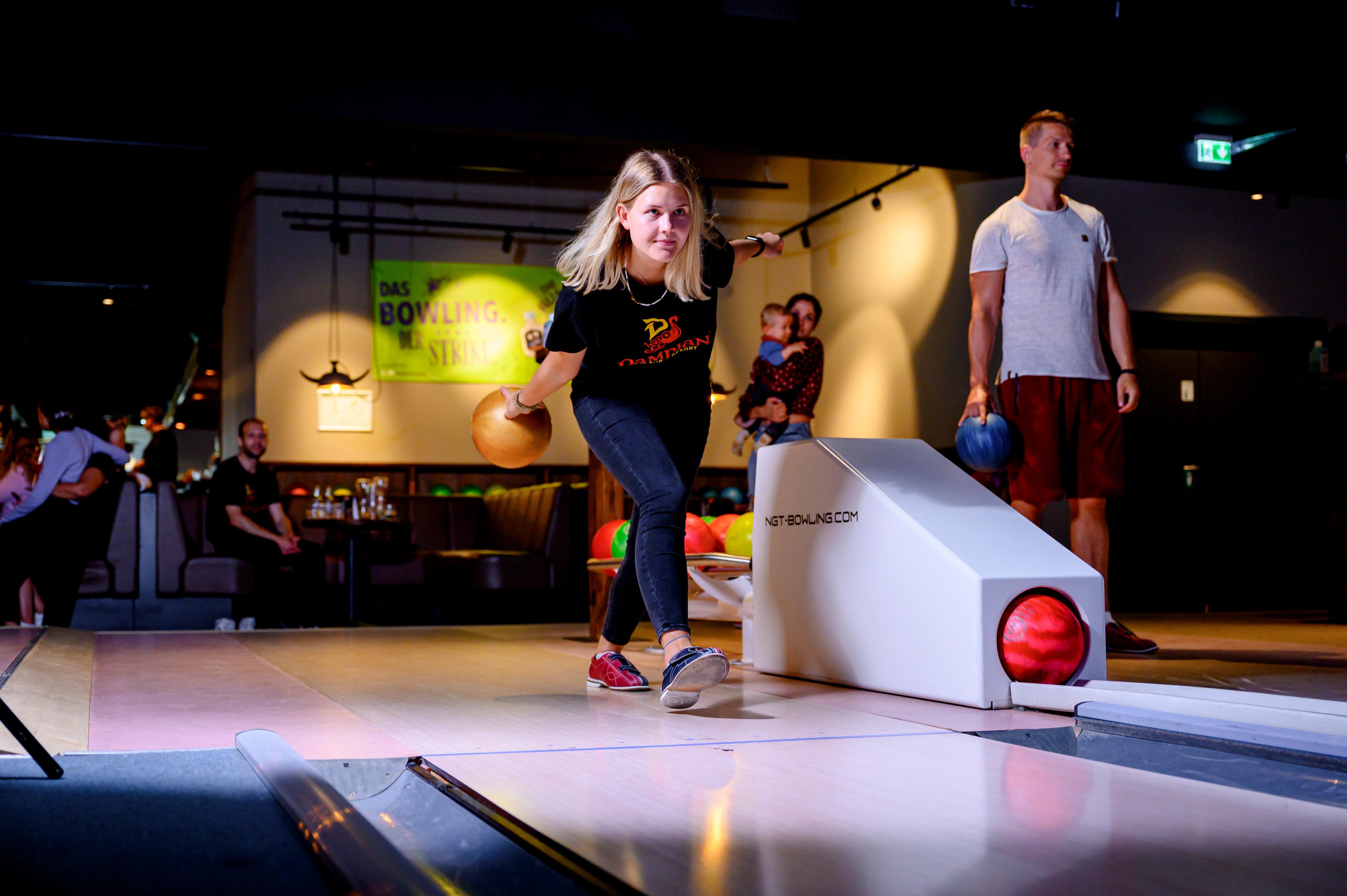 Eine Frau wirft eine Bowlingkugel in einer Bowlinghalle, während ein Mann im Hintergrund steht.