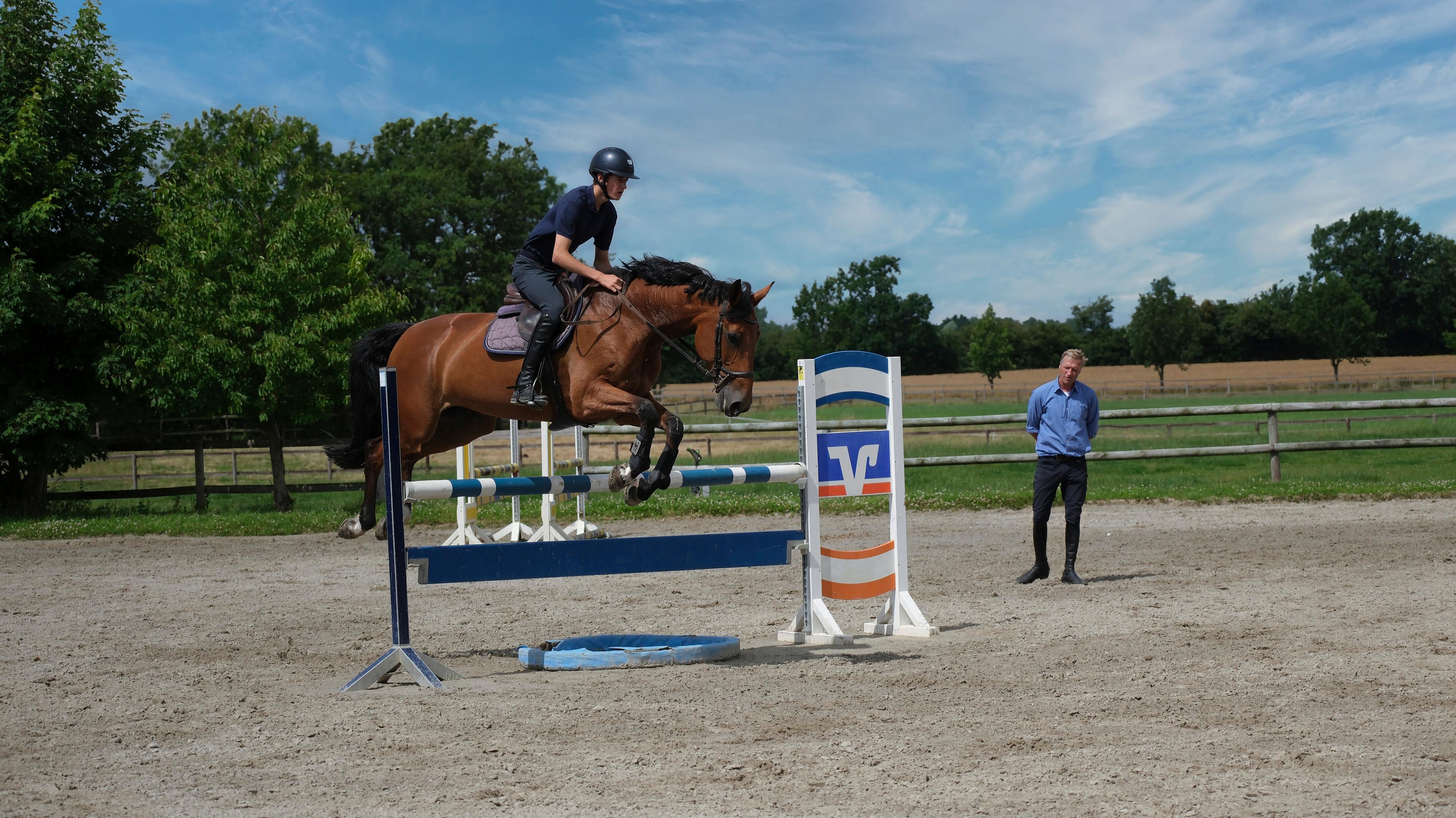 Ein Reiter springt mit einem braunen Pferd über ein Hindernis auf einem Außenplatz, während ein Mann zuschaut.