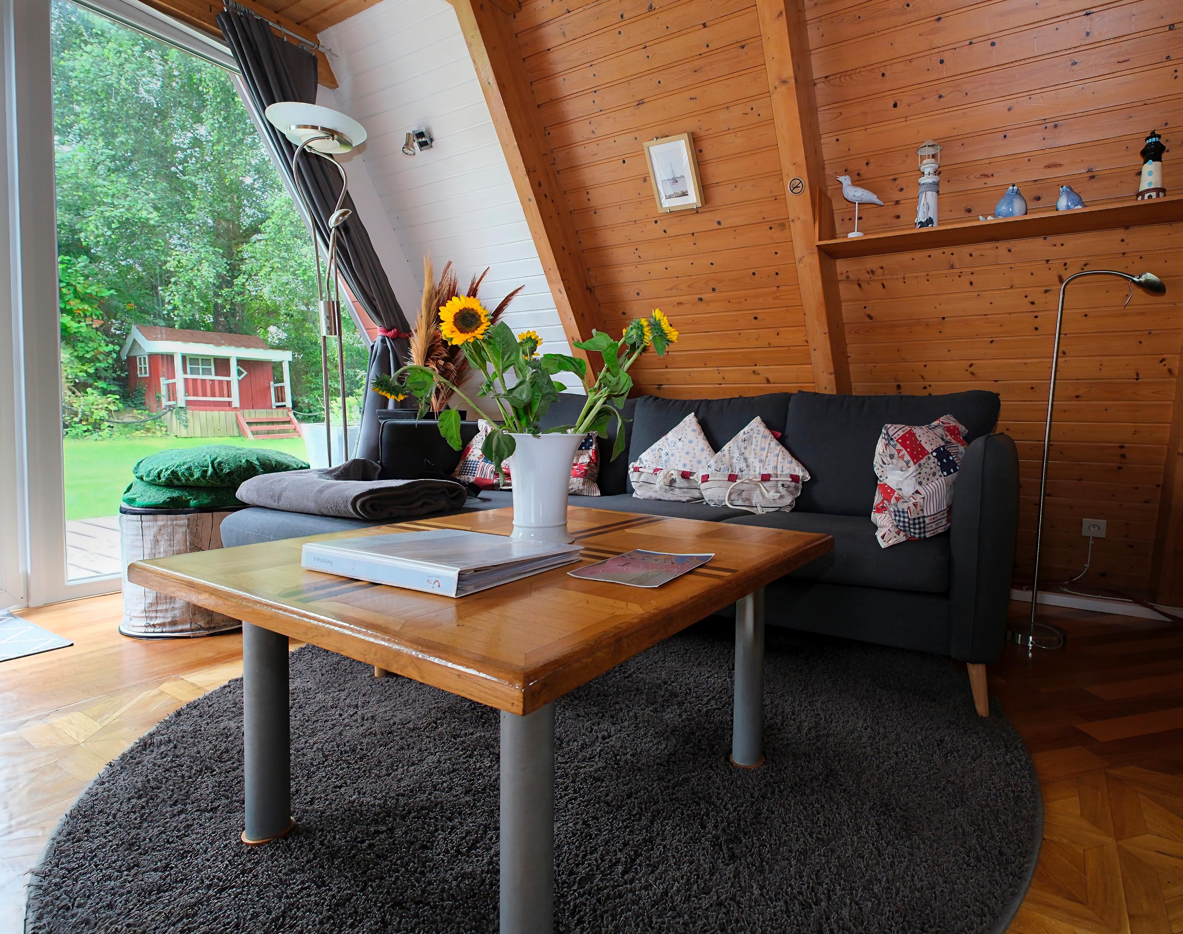 Ein gemütliches Wohnzimmer mit Holzwänden, einem grauen Sofa, einem Holztisch mit Sonnenblumen und Blick auf einen Garten mit rotem Gartenhaus.