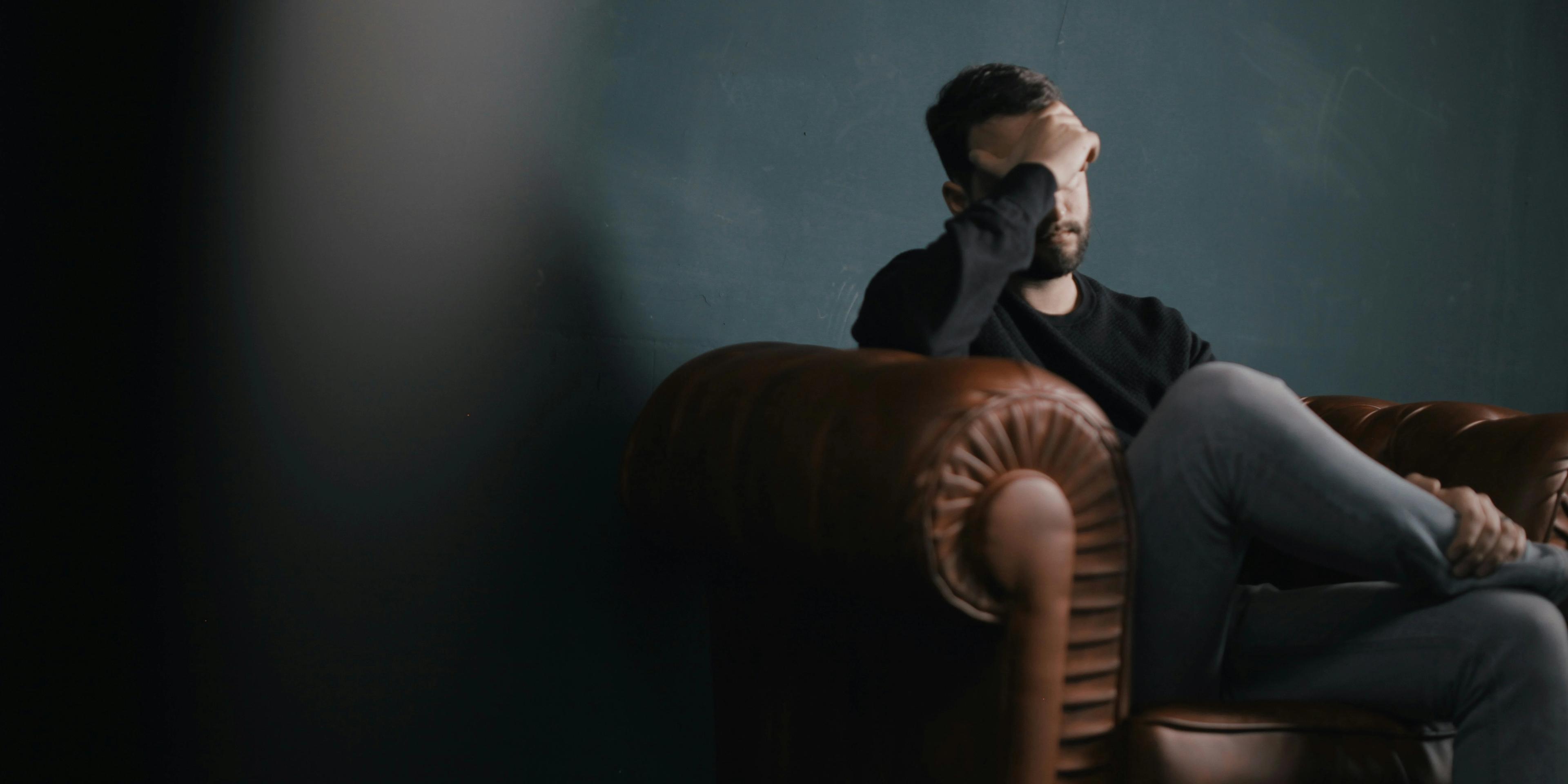 A person sitting on a couch, leaning forward with head bowed, in a dimly lit room, conveying a sense of stress or sadness.