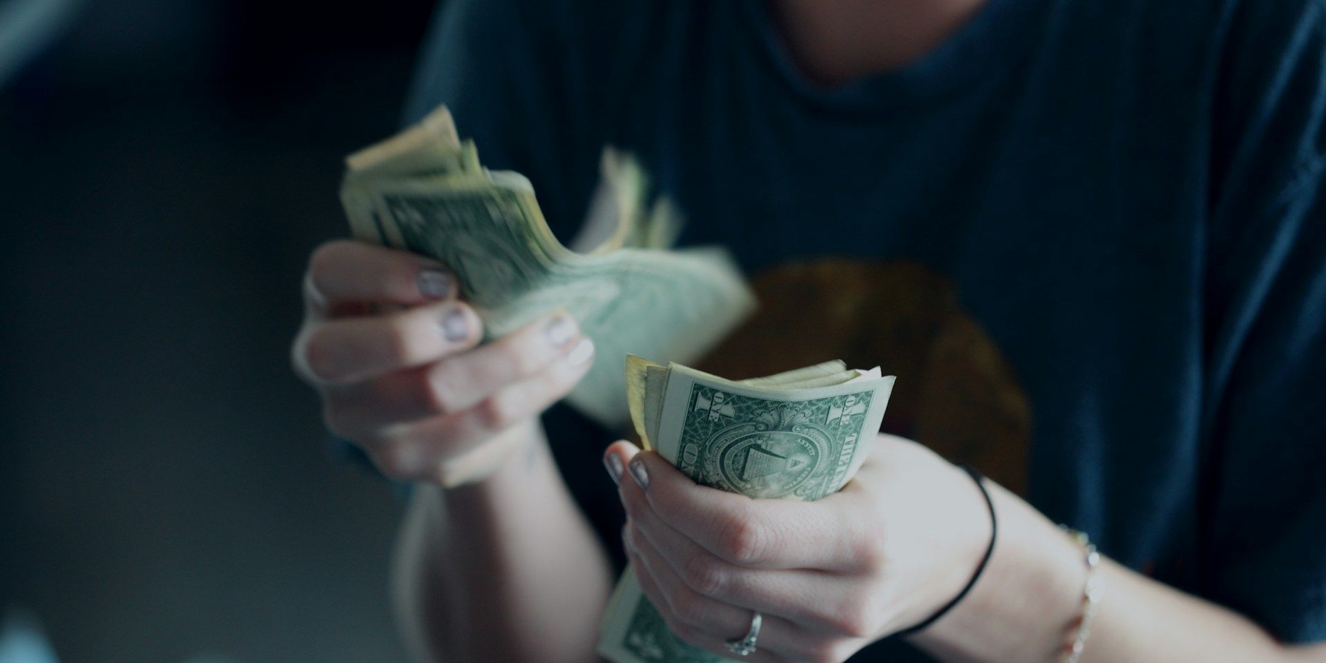 Person counting dollar bills with both hands, sitting at a table with a cup nearby.