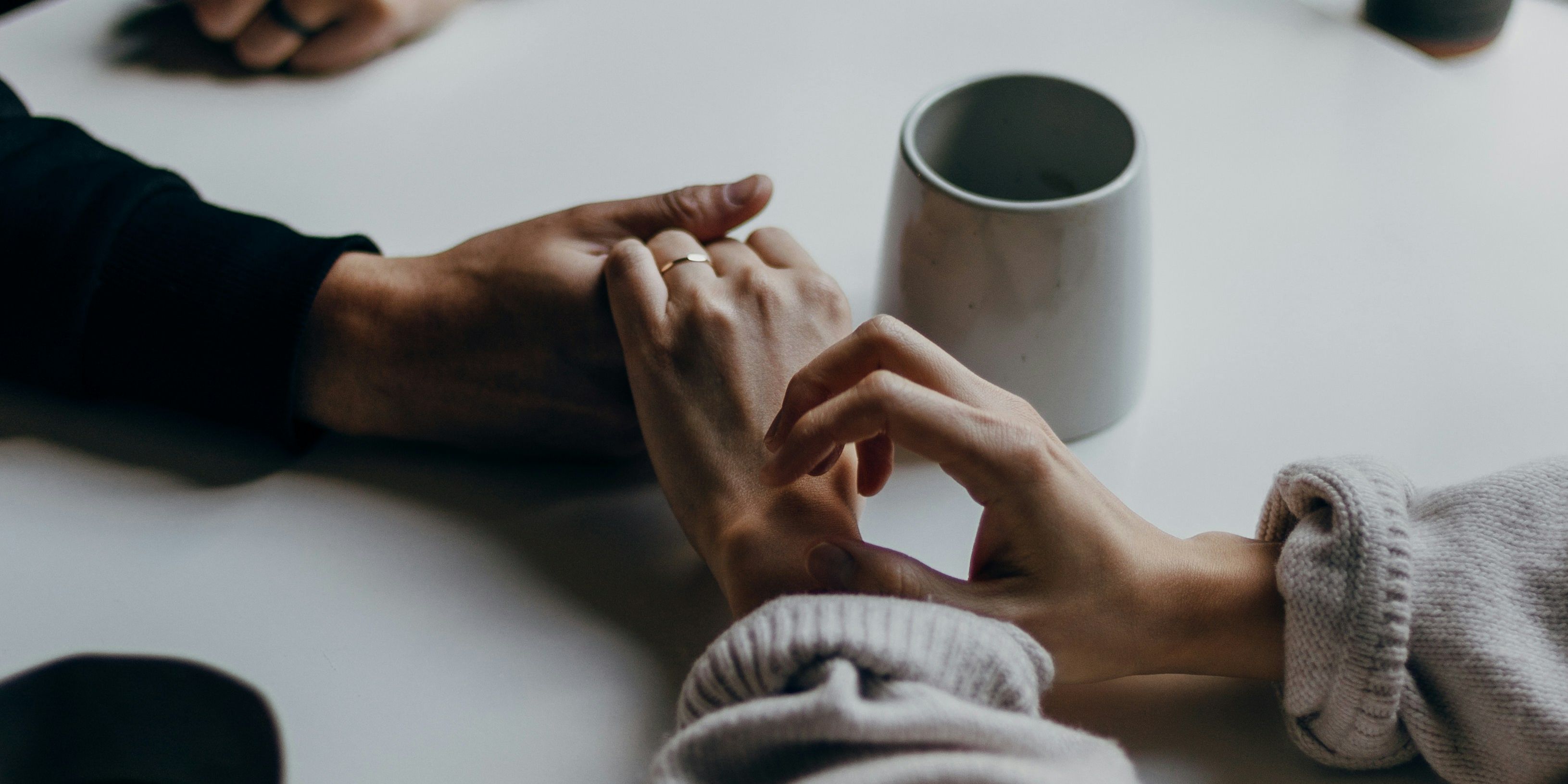 Here’s an appropriate alt text for the image you just provided: **"Close-up of two people holding hands across a white table, coffee mugs nearby, soft natural light coming from a window."**
