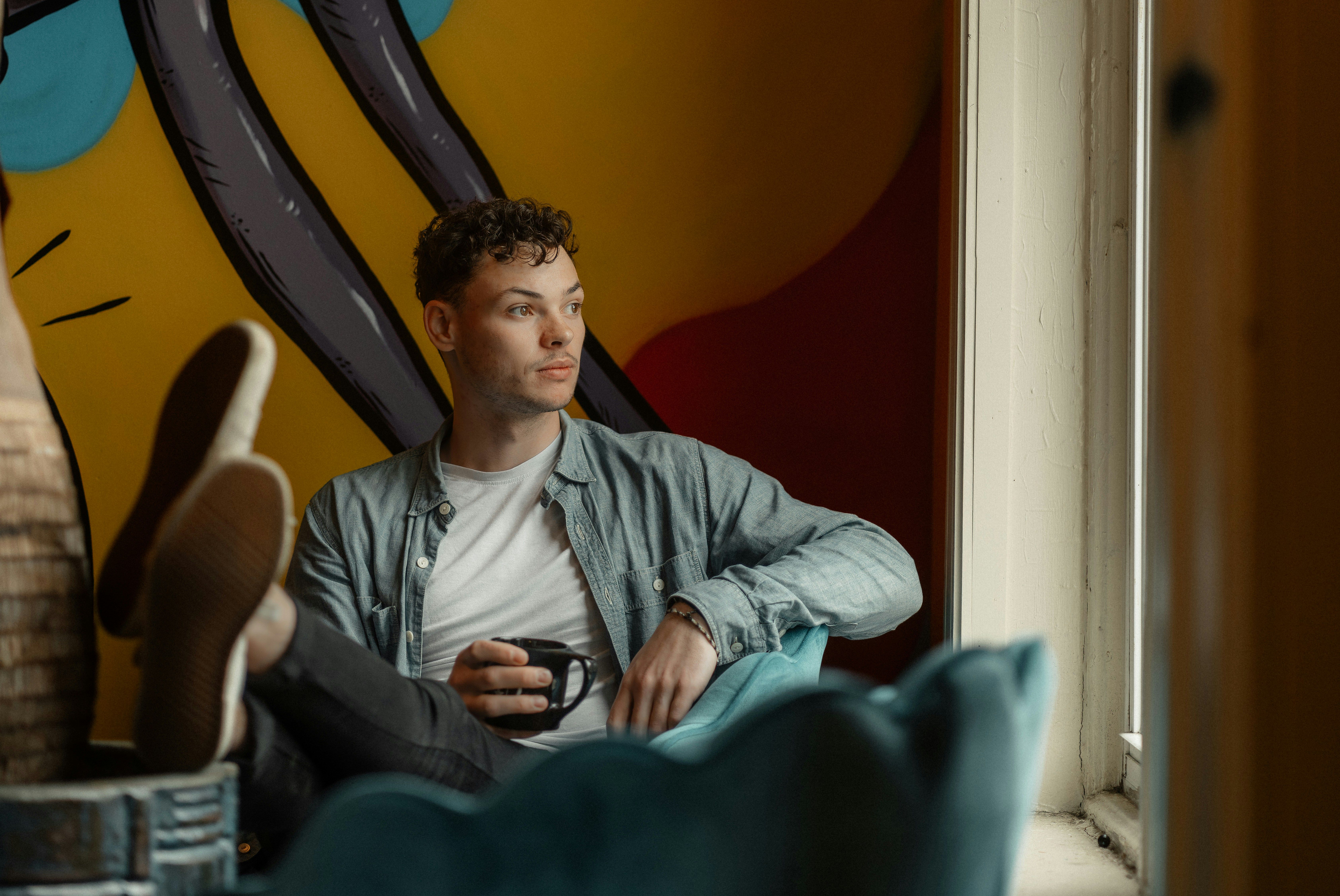 Young man sitting indoors, looking thoughtfully out of a window while holding a coffee mug.