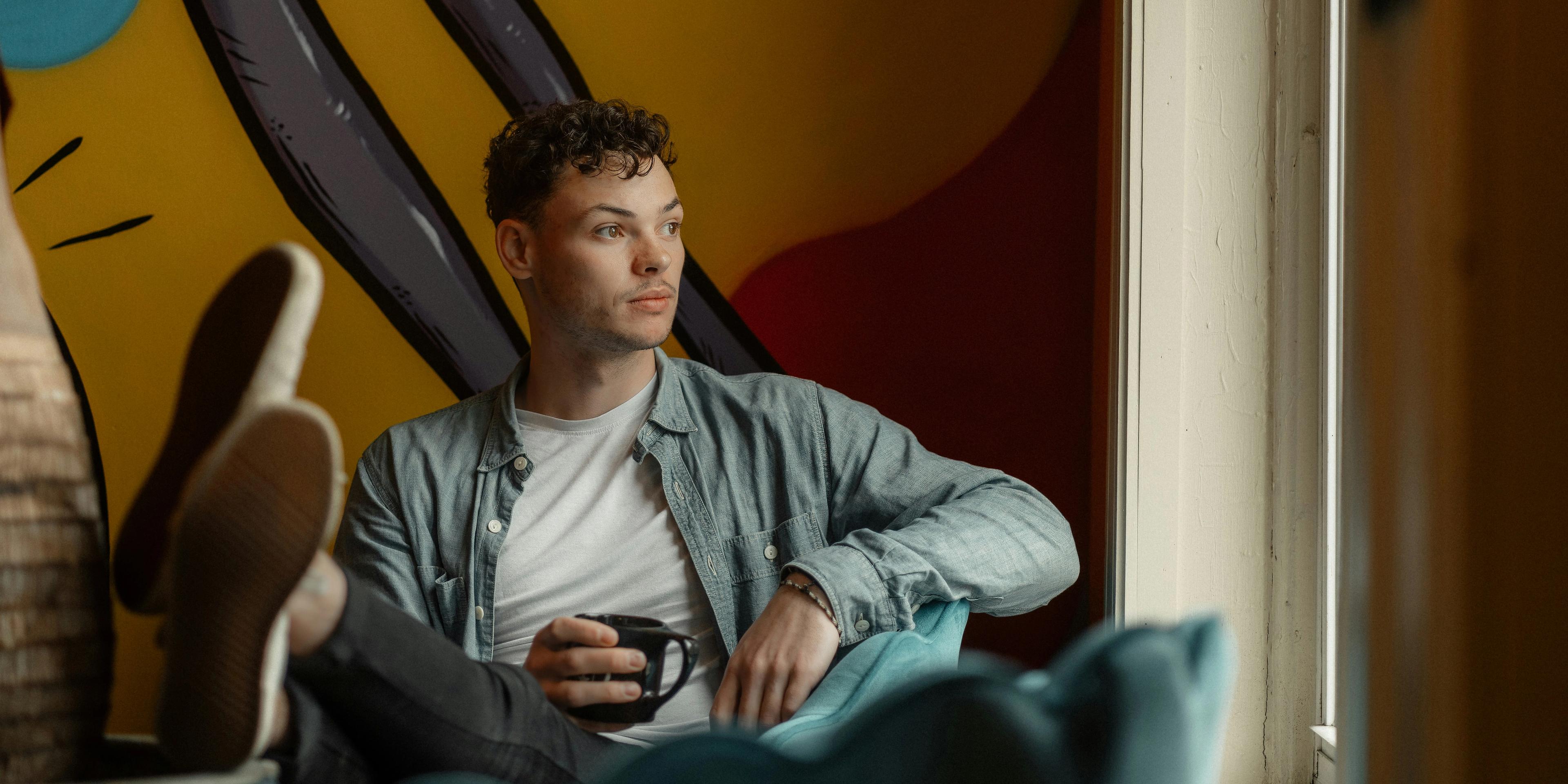 Young man sitting indoors, looking thoughtfully out of a window while holding a coffee mug.