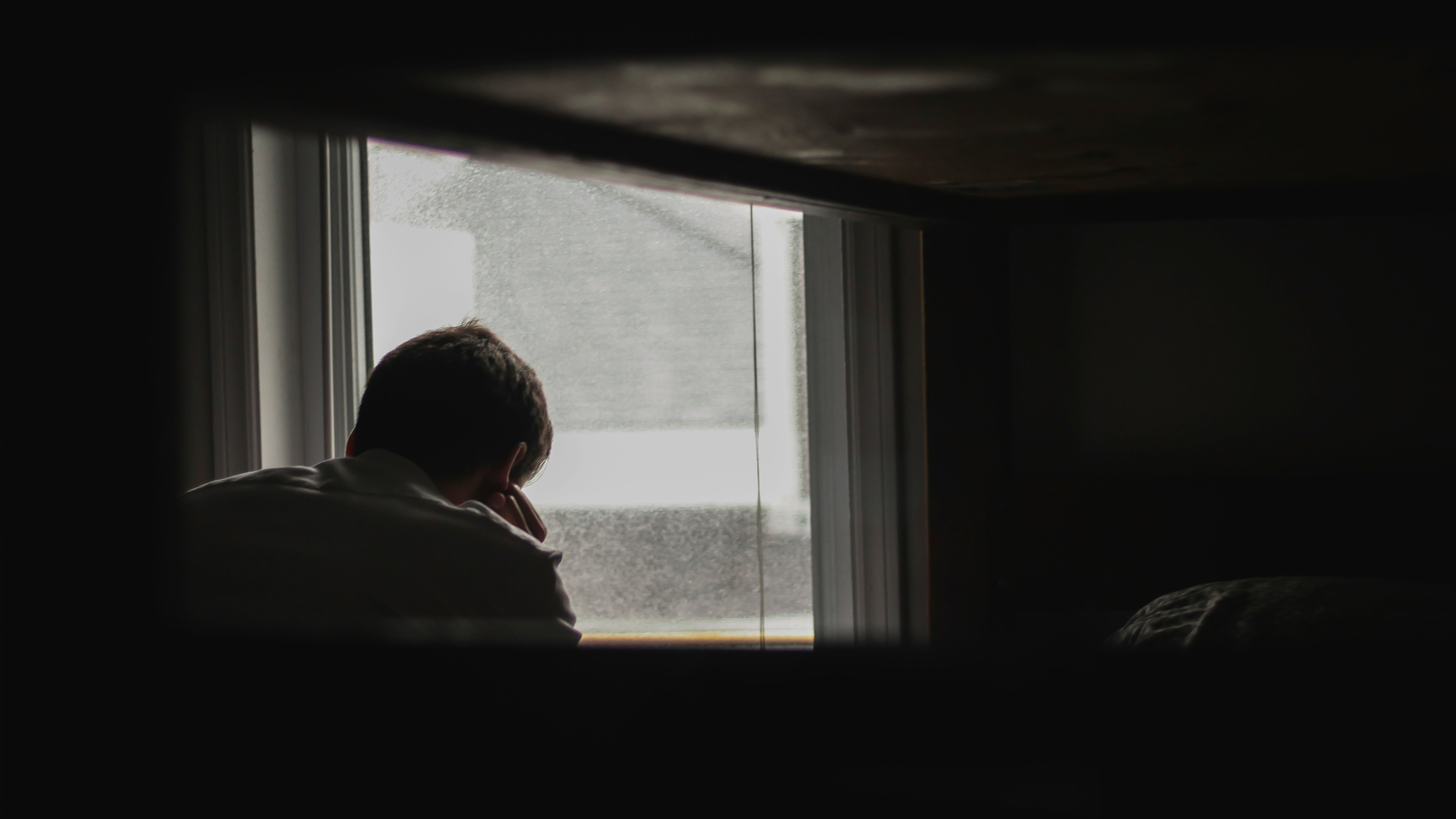 Person sitting by a window, looking outside with their back to the camera, framed by a dark interior.