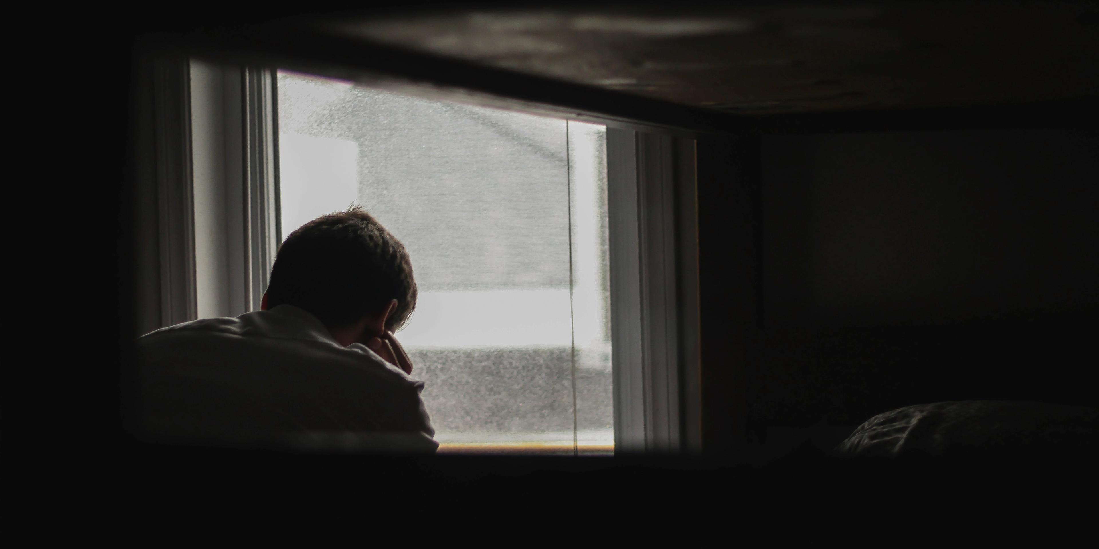 Person sitting by a window, looking outside with their back to the camera, framed by a dark interior.