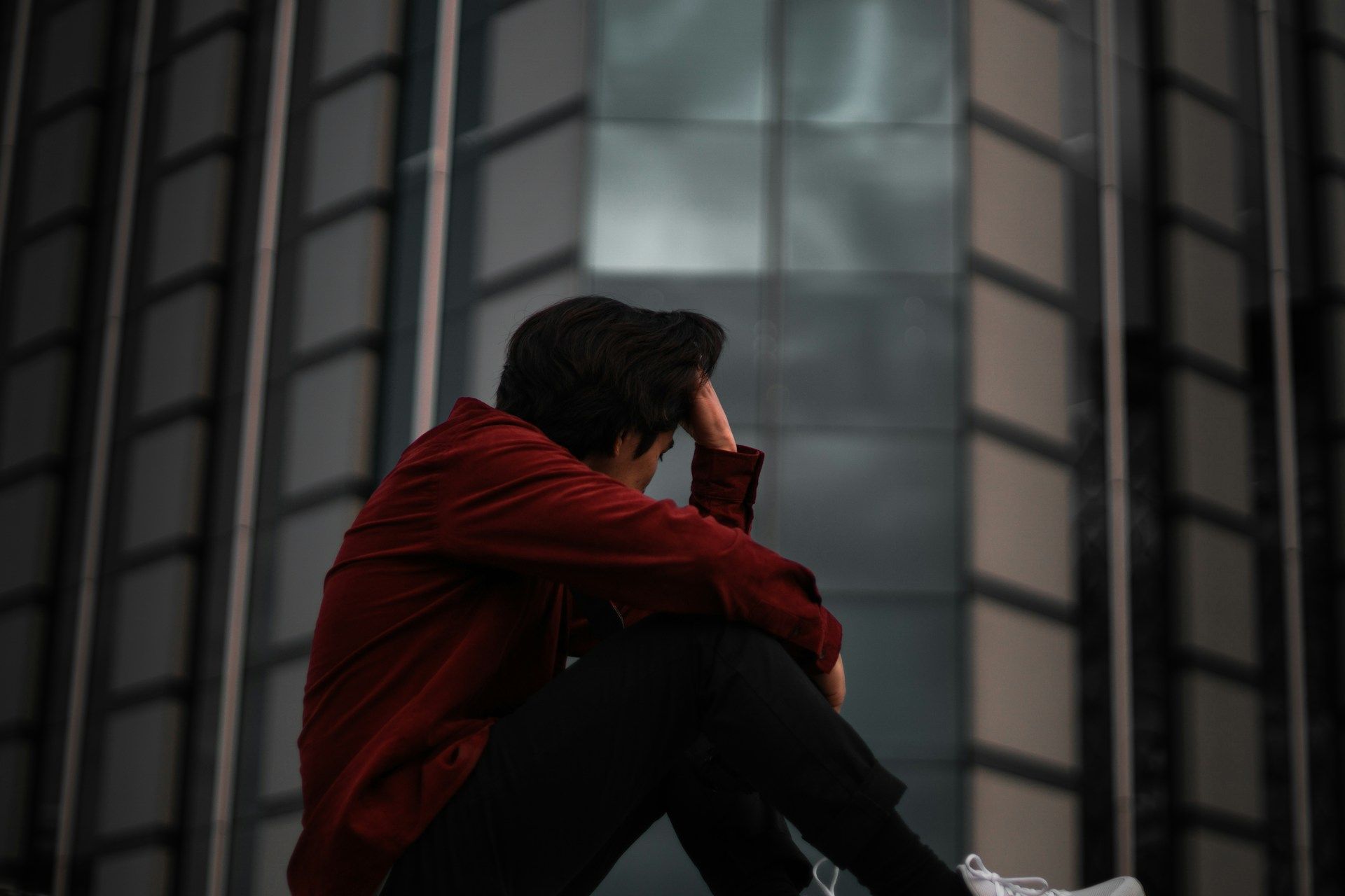 Person sitting alone and appearing distressed outside a modern building, illustrating the emotional impact of a gambling problem