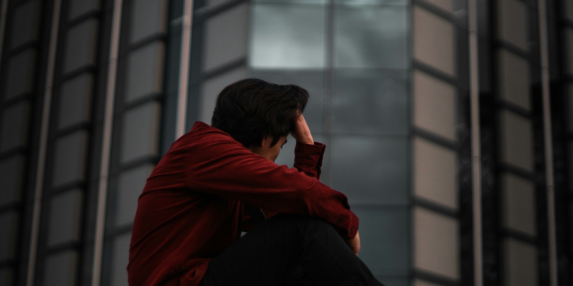 Person sitting alone and appearing distressed outside a modern building, illustrating the emotional impact of a gambling problem