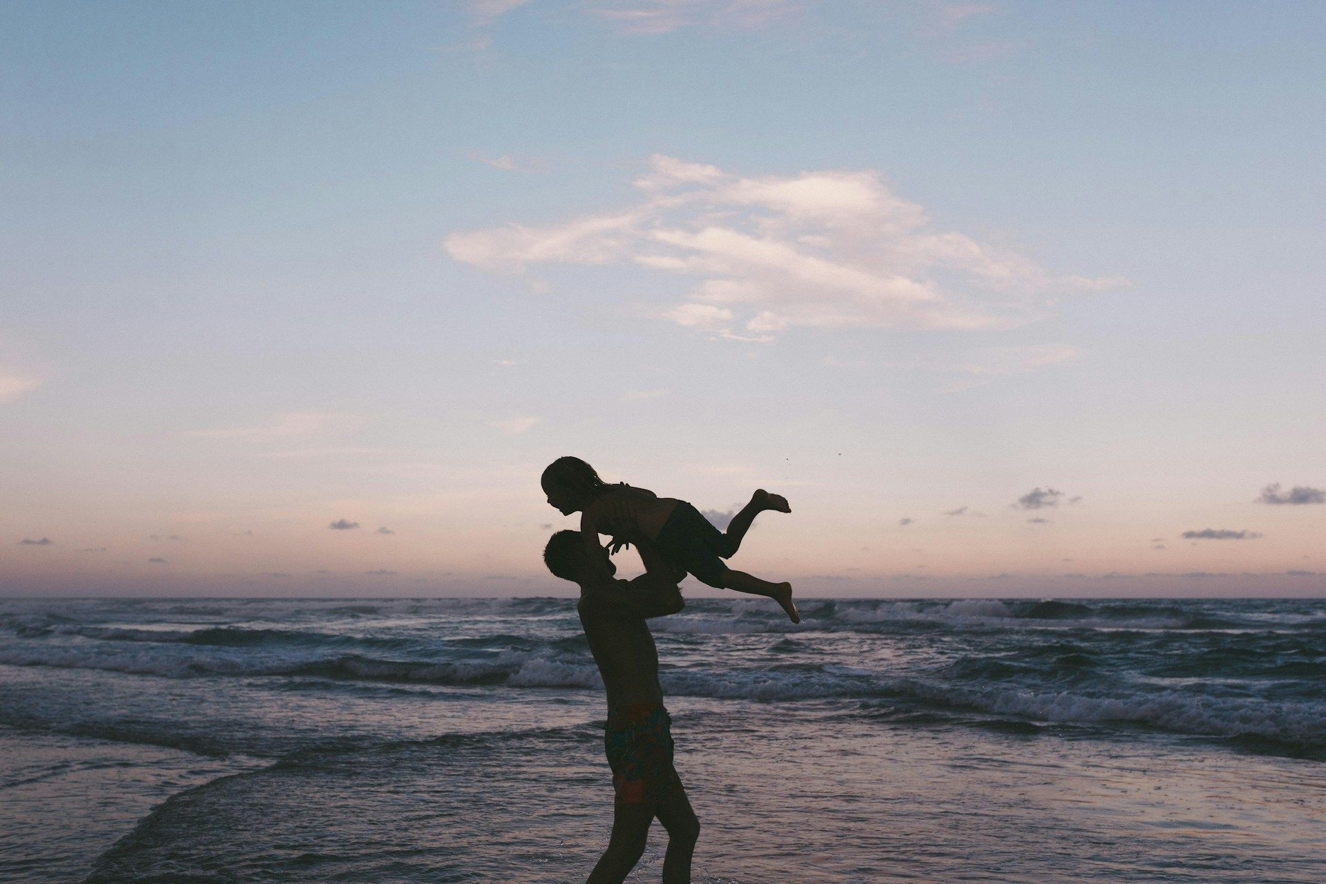 Silhouette of a parent lifting a child above their head at the beach during sunset