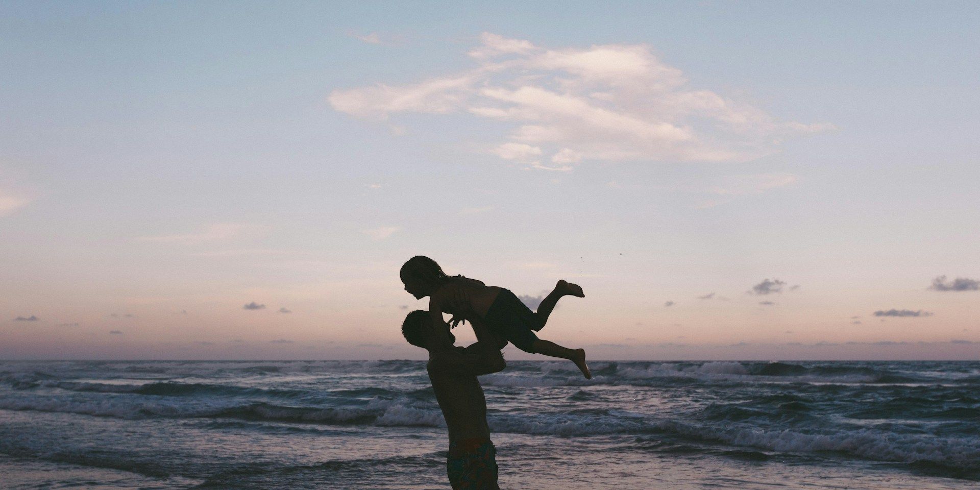 Silhouette of a parent lifting a child above their head at the beach during sunset