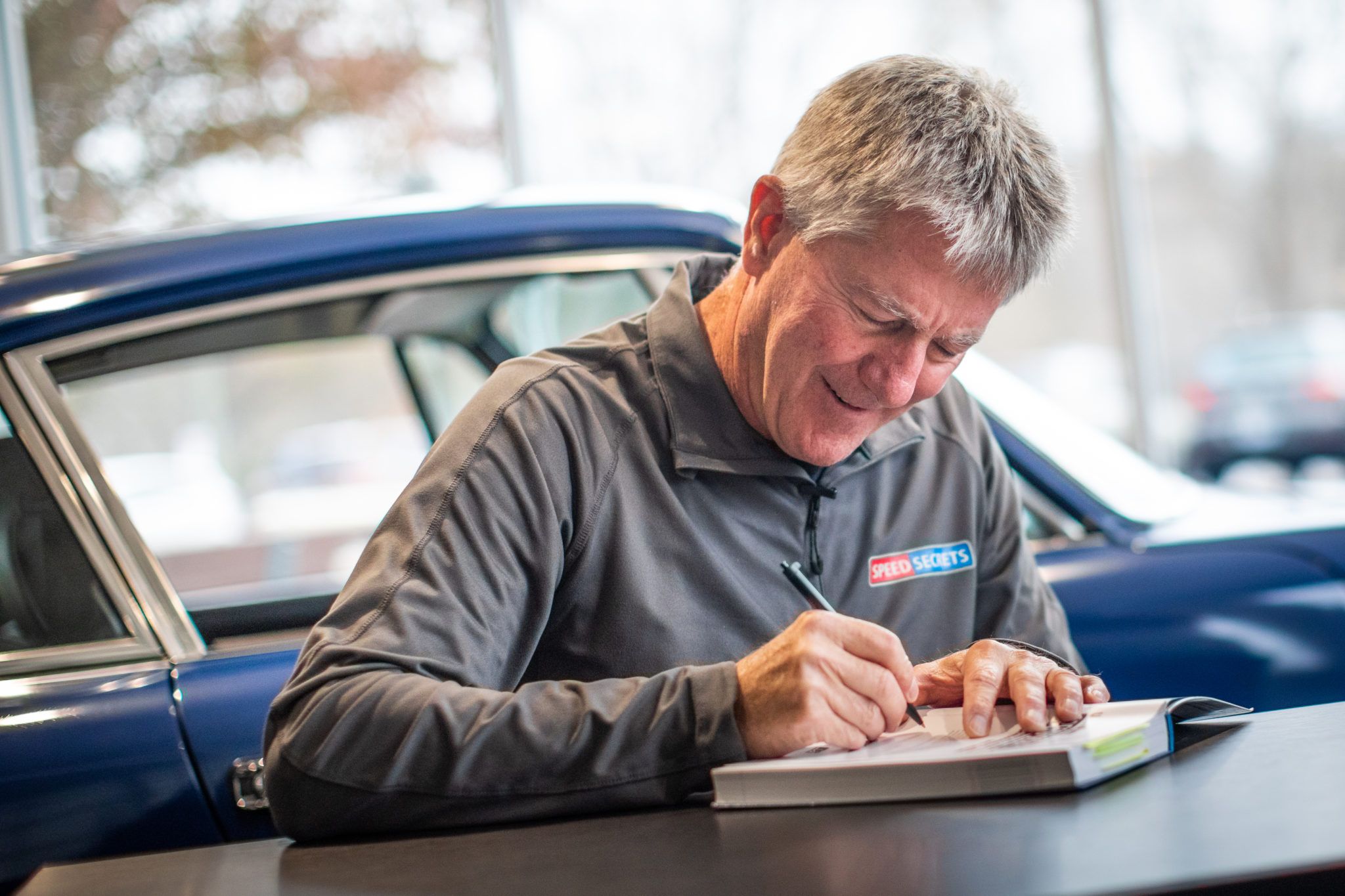 Ross Bentley smiling, signing a SpeedSecrets book at a table, in front of a classic sports car