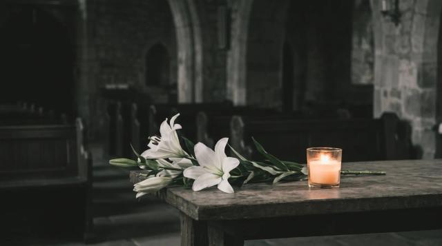White lilies and a lit memorial candle in a softly lit chapel, representing a dignified burial or cremation.
