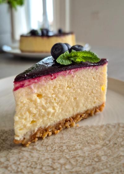 Cremiger New York Cheesecake mit glattem Heidelbeerspiegel und nussigem Mandelboden auf einem rustikalen Kuchenteller.