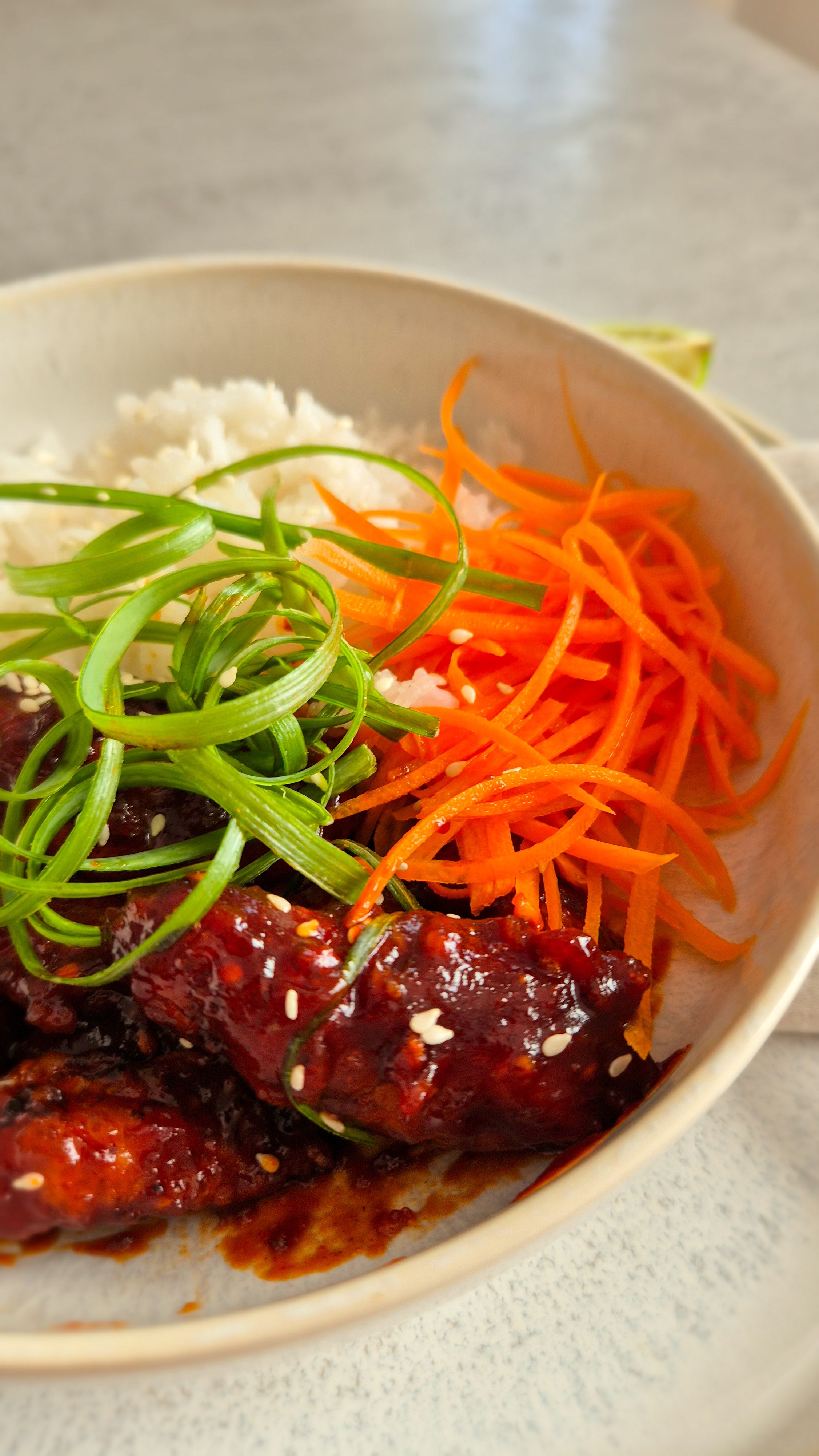 Koreanisches Fried Chicken - Yangnyeom - mit Frühlingszwiebeln und Sesam zu Reis und Karotten in einer Schale serviert.