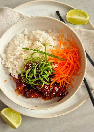Koreanisches Fried Chicken - Yangnyeom - mit Frühlingszwiebeln und Sesam zu Reis und Karotten in einer Schale serviert.