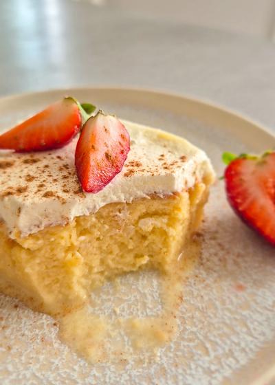 Ein Stück saftiger Tres Leches Kuchen (3 Milch Kuchen) aus Lateinamerika mit einer Sahnehaube und frischen Erdbeeren auf einem Teller serviert.