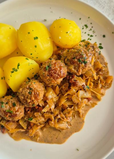Hackbällchen mit gedünstetem Weißkohl in cremiger Sauce und Salzkartoffeln auf einem Teller.