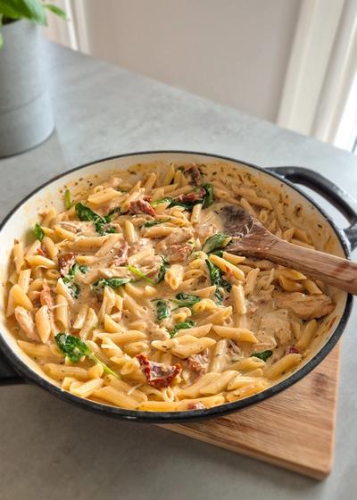Cremige Marry Me Chicken Pasta mit sahniger Sauce, getrockneten Tomaten und Blattspinat in einer Pfanne auf einem Holzbrett.