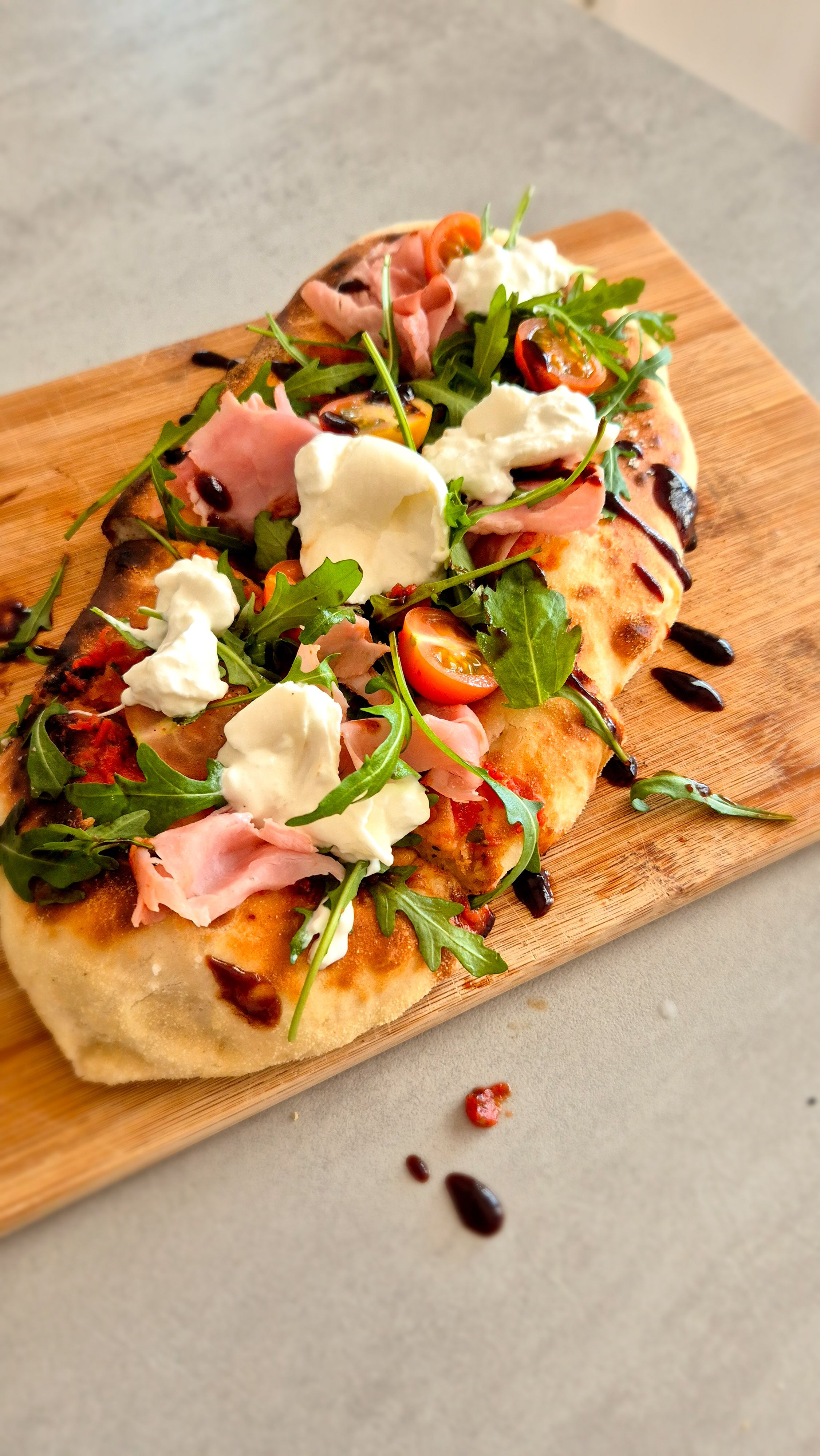 Rustikale Pinsa mit Burrata, Prosciutto und Rucola auf einem Brett.