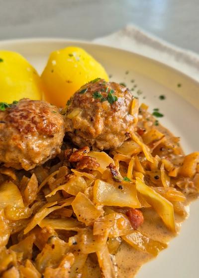 Hackbällchen mit gedünstetem Weißkohl in cremiger Sauce und Salzkartoffeln auf einem Teller.