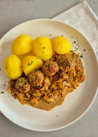 Hackbällchen mit gedünstetem Weißkohl in cremiger Sauce und Salzkartoffeln auf einem Teller.