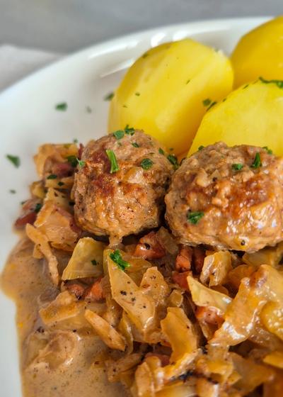 Hackbällchen mit gedünstetem Weißkohl in cremiger Sauce und Salzkartoffeln auf einem Teller.