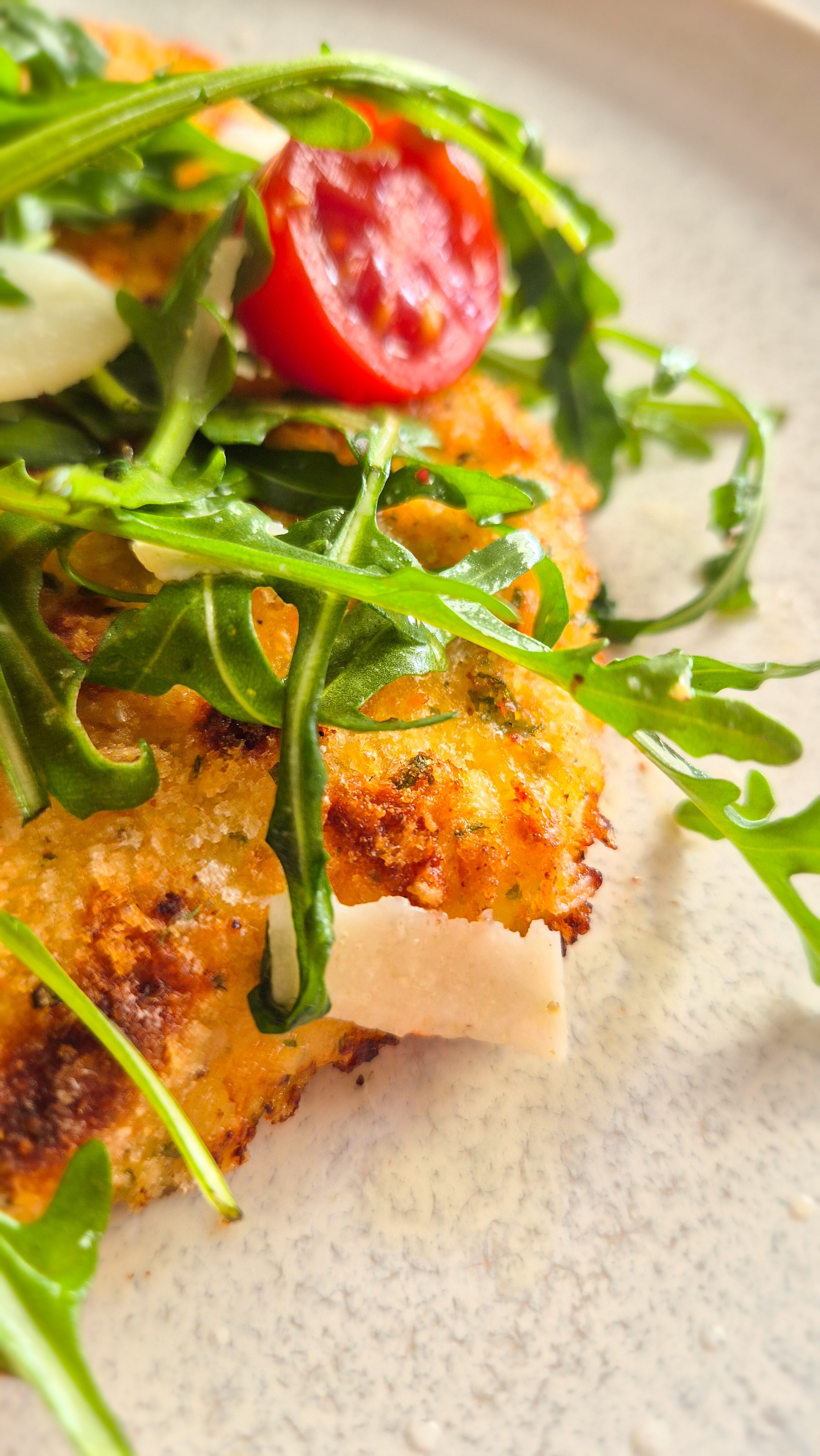Ein Teller mit knusprig gebackenem Chicken Milanese, belegt mit frischem Rucola, Cherrytomaten und Parmesan-Hobeln. Nahaufnahme.