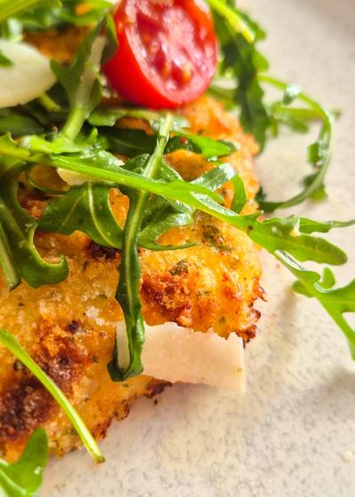 Ein Teller mit knusprig gebackenem Chicken Milanese, belegt mit frischem Rucola, Cherrytomaten und Parmesan-Hobeln. Nahaufnahme.