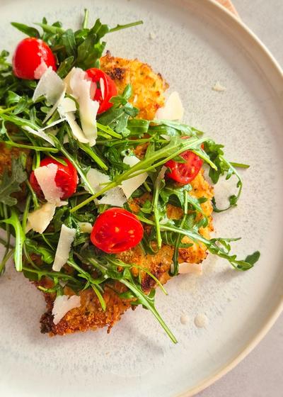 Ein Teller mit knusprig gebackenem Chicken Milanese, belegt mit frischem Rucola, Cherrytomaten und Parmesan-Hobeln.
