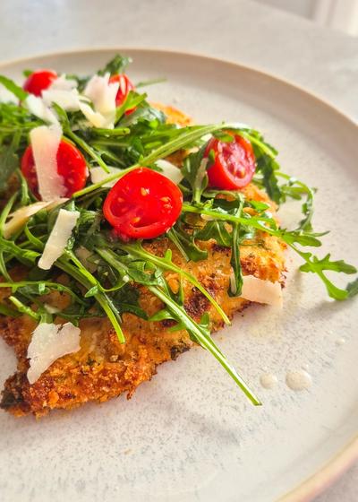 Ein Teller mit knusprig gebackenem Chicken Milanese, belegt mit frischem Rucola, Cherrytomaten und Parmesan-Hobeln.